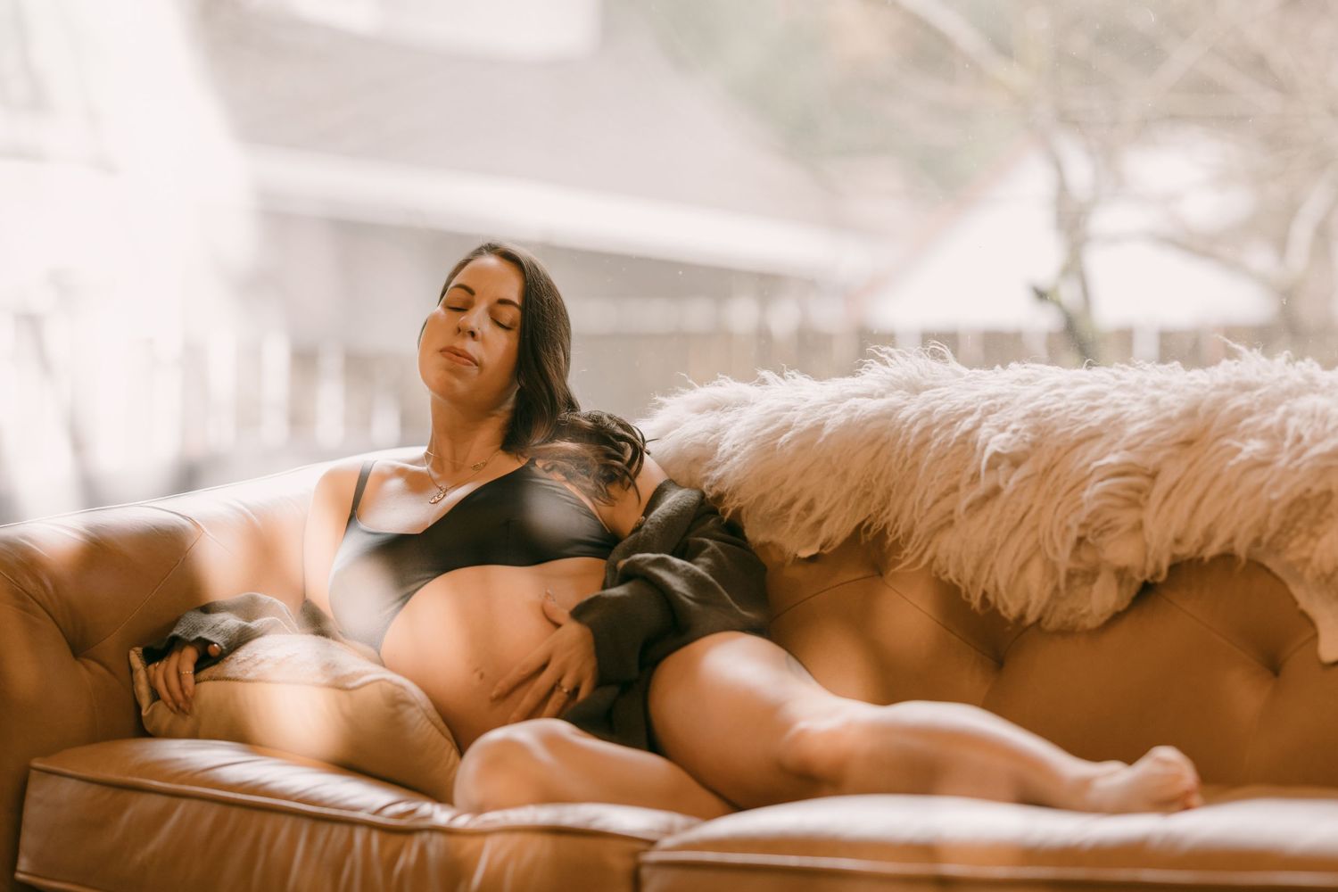 Pregnant woman in black lingerie reclines on leather sofa with fluffy fur throw in natural light.