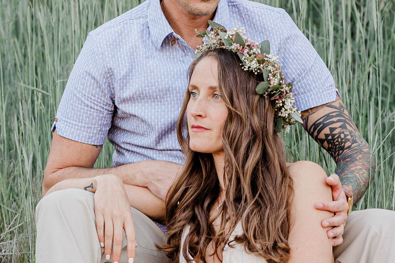 Couple sits in tall grass, woman with floral crown leans against tattooed partner.