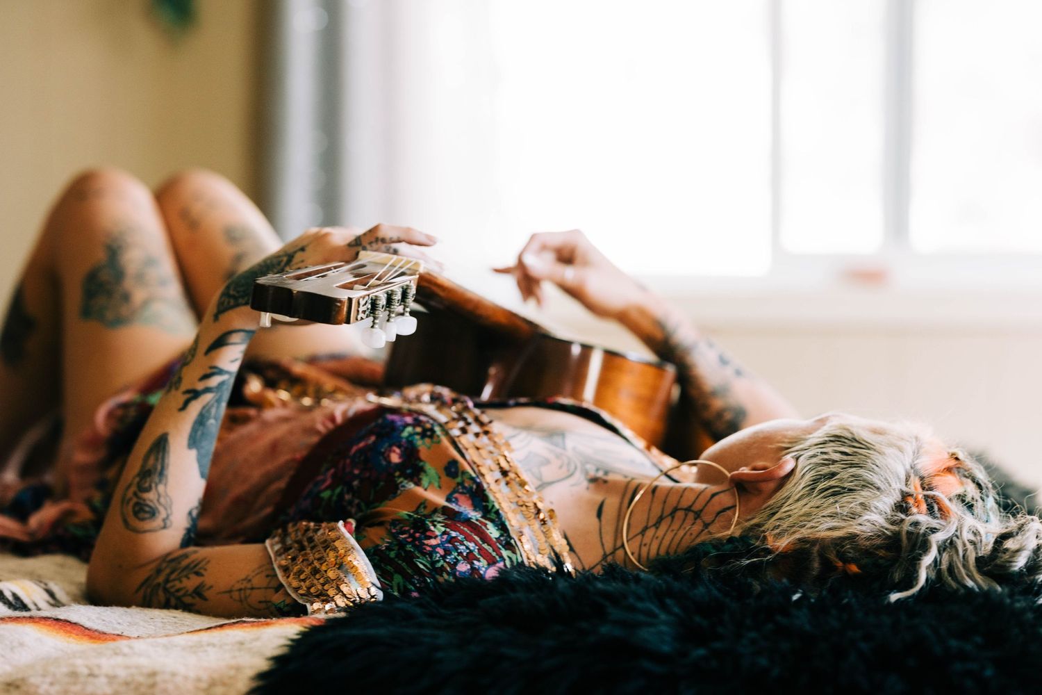 Heavily tattooed woman lying on a fur rug holding a guitar wearing a bohemian outfit.