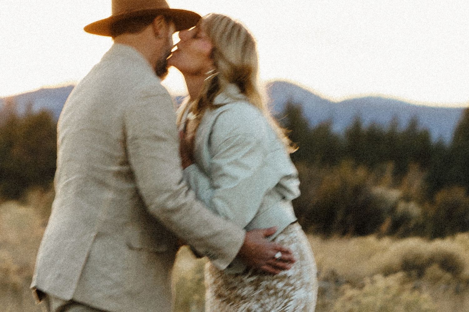 Newlyweds share a romantic kiss in a sunlit meadow with mountains in the background.