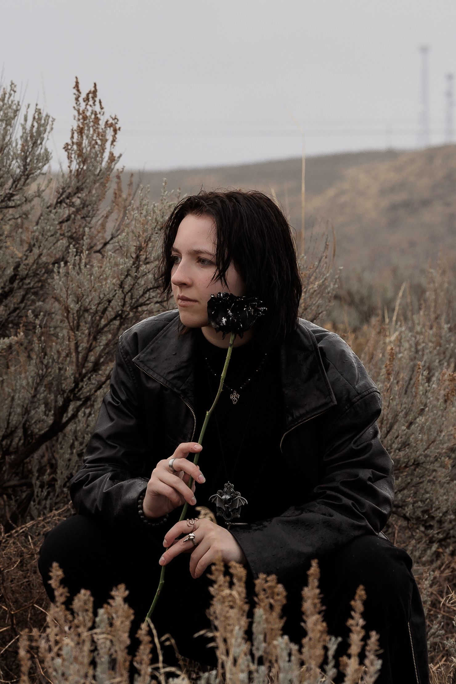 Dark-haired person in black jacket holding a dried black flower while sitting in desert shrubs.