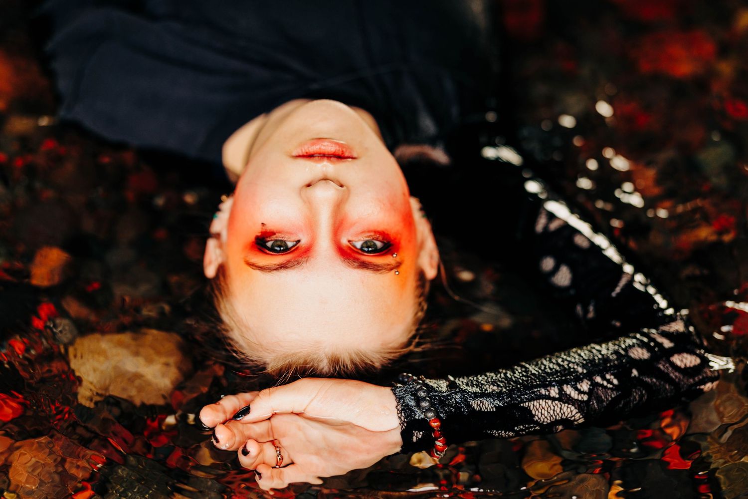 Woman with vivid red eye makeup lies upside down in shallow water among colorful rocks.