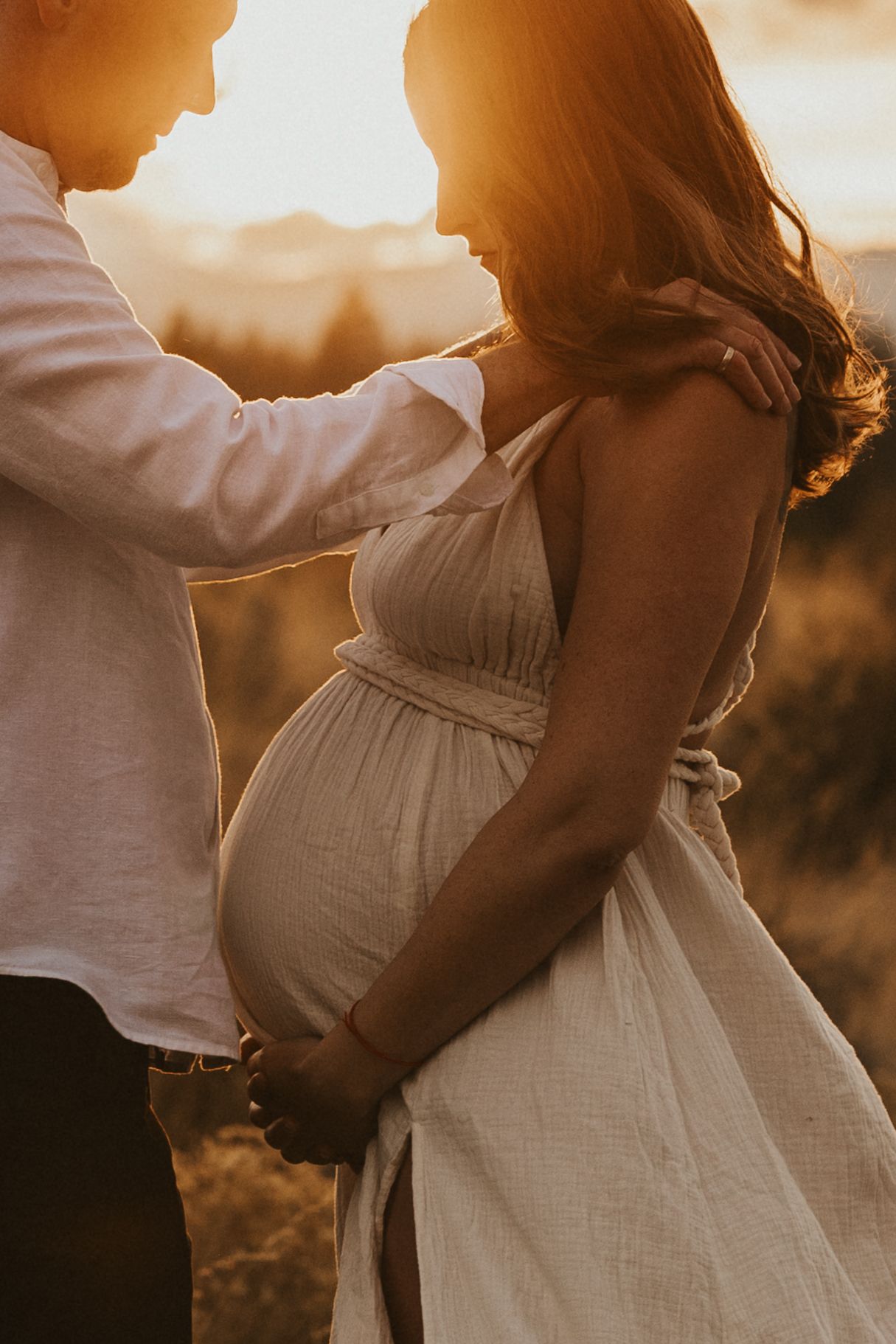 Couple embracing during golden hour maternity shoot in an outdoor field at sunset.