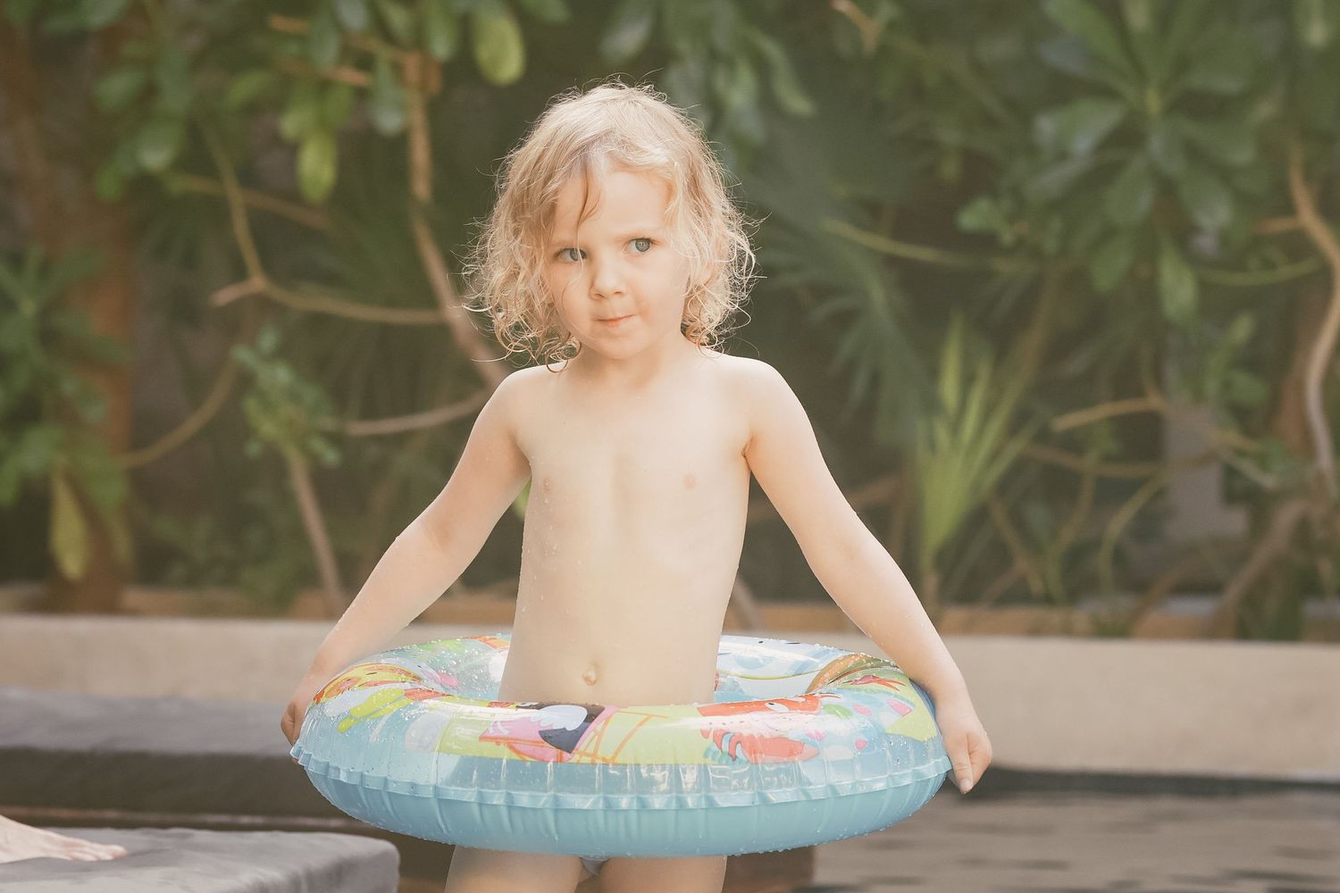 Young blonde child standing outdoors holding a colorful inflatable swim ring near a pool.