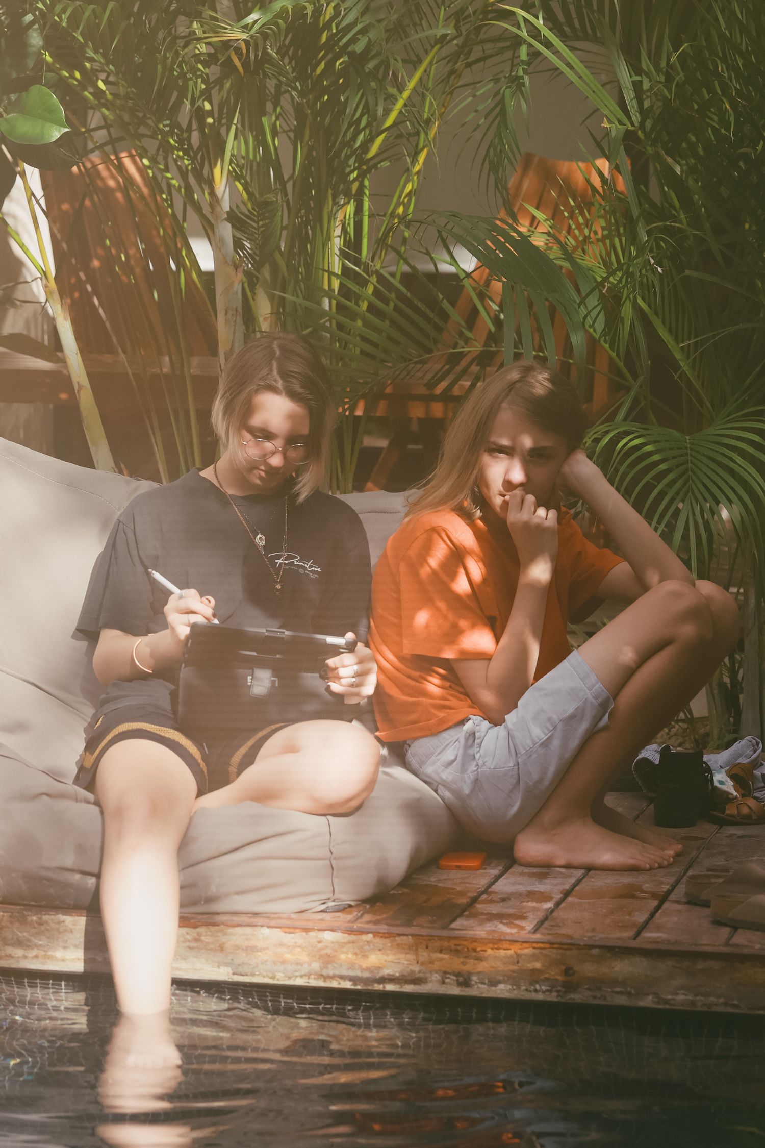 Two teenagers sitting on a poolside bean bag reading together surrounded by tropical green plants.
