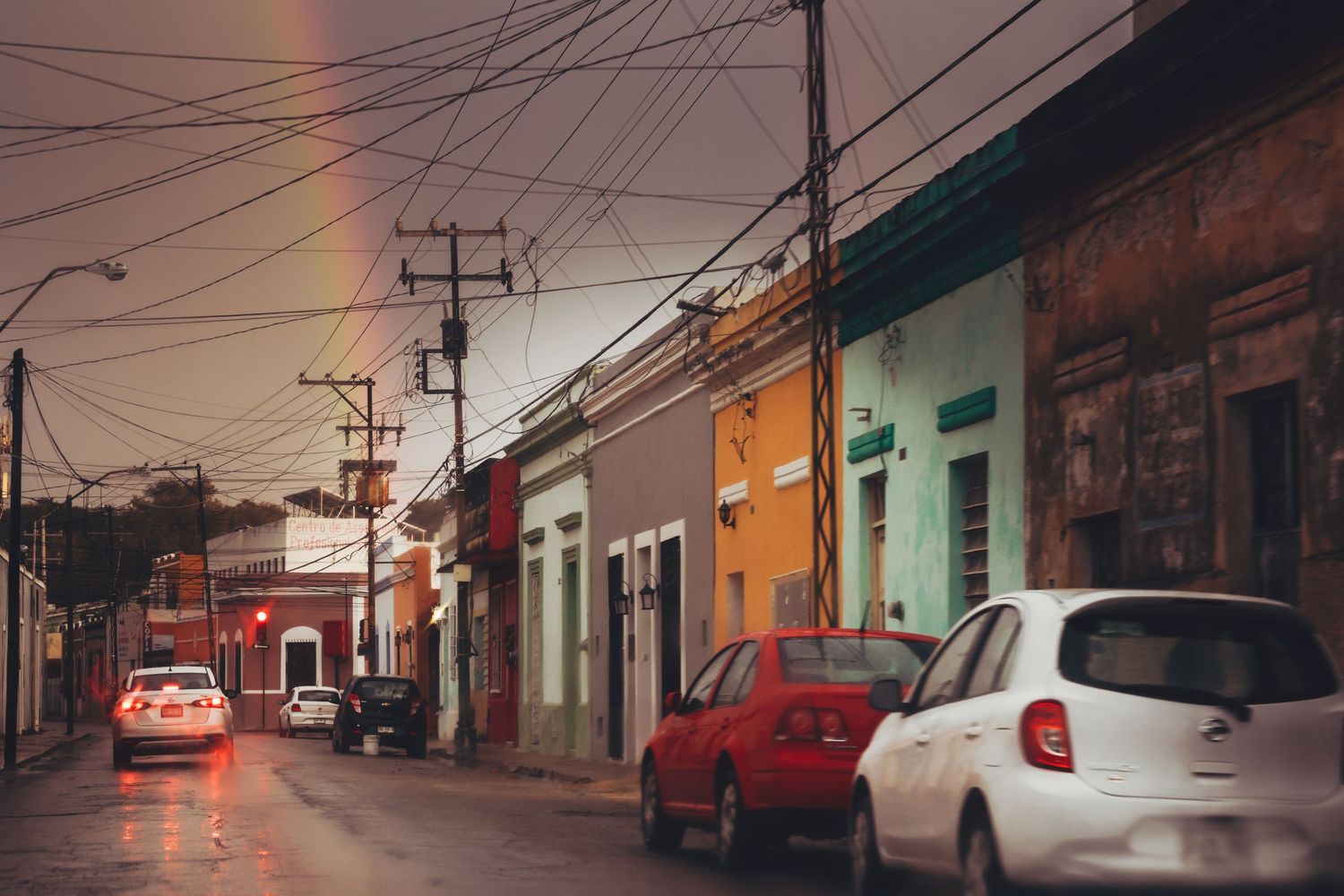 Rainy evening street scene with colorful colonial buildings, parked cars, and a rainbow in the sky.
