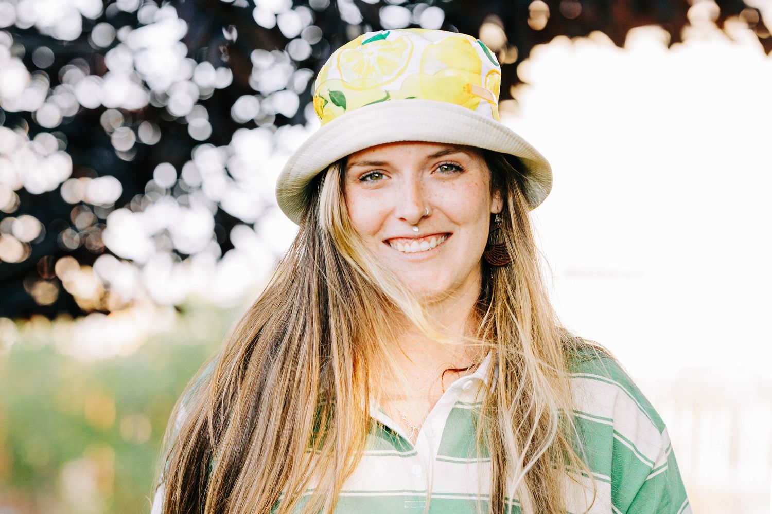 Smiling young woman with long brown hair wearing plaid shirt and yellow bucket hat.