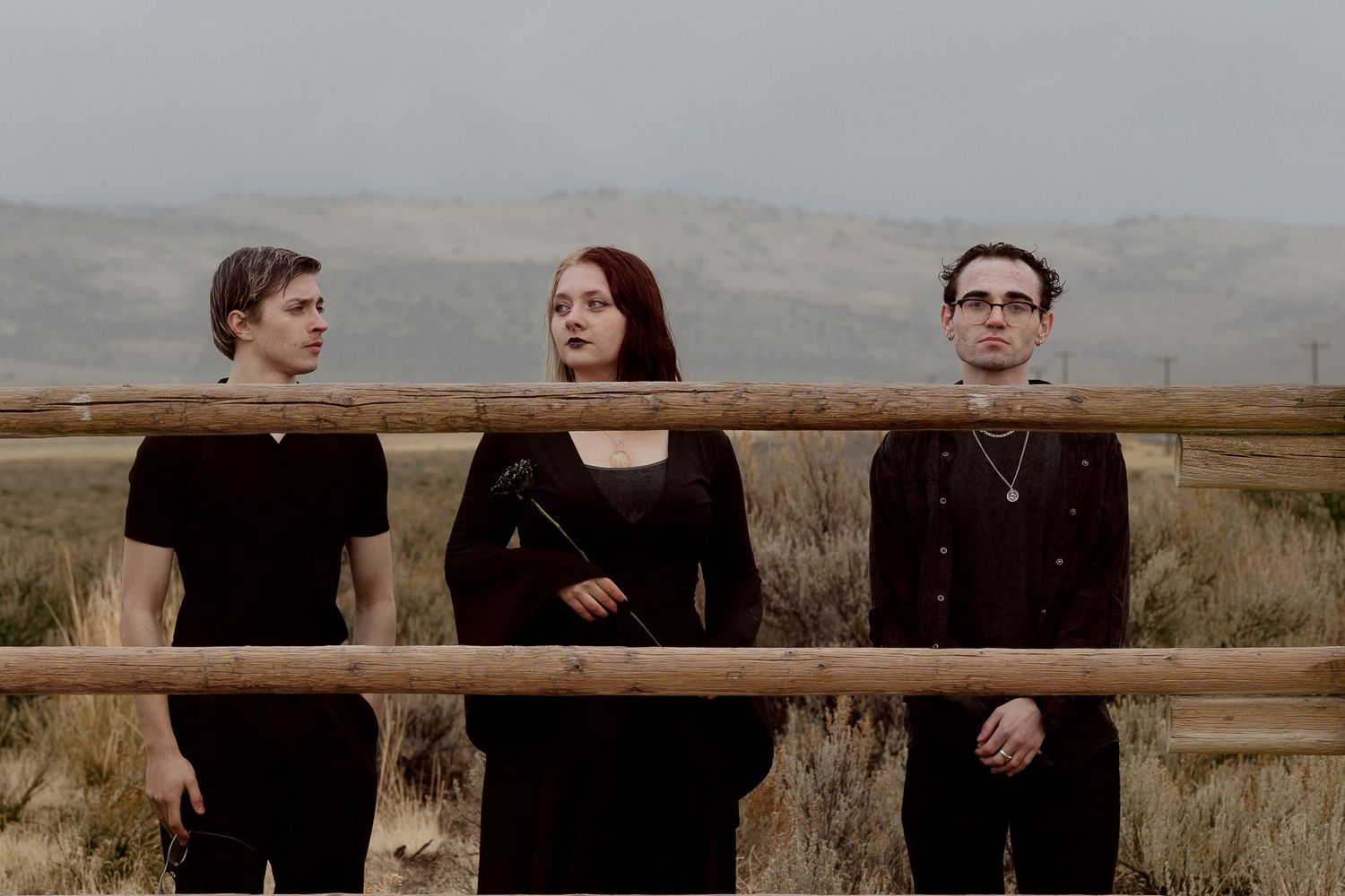 Three people in black gothic attire pose by a wooden fence with misty mountains and dry grasslands behind them.