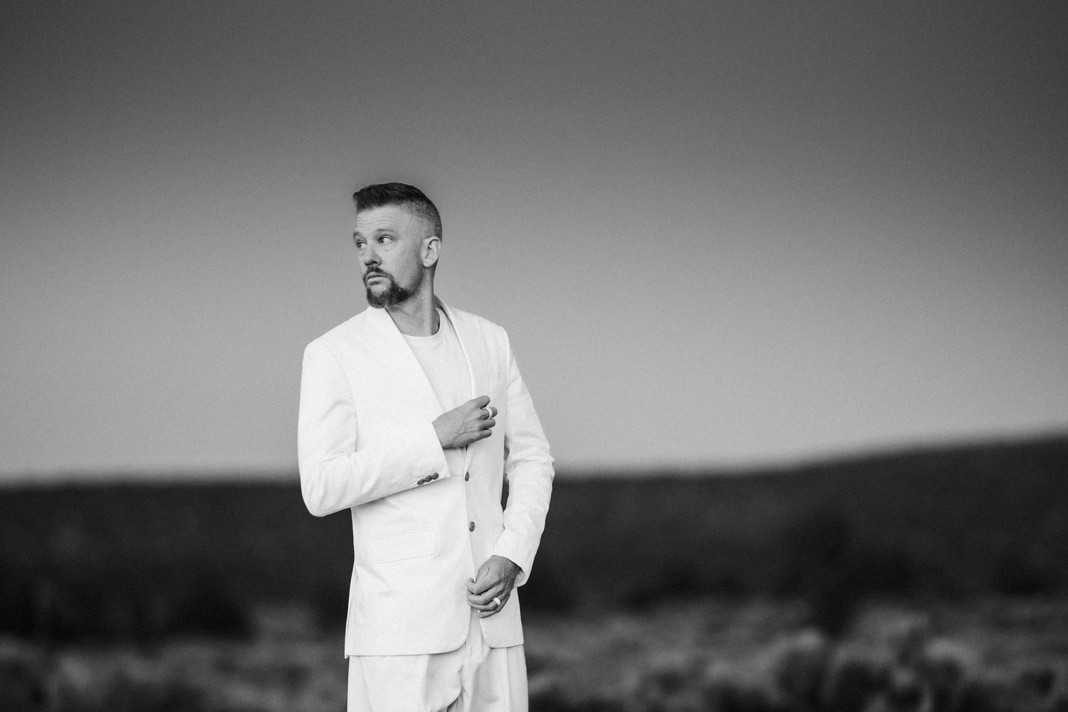 Black and white photo of bearded man in white suit standing in open desert field.