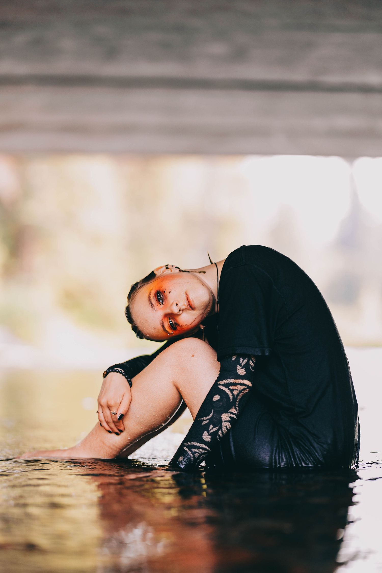 Smiling woman with orange eye makeup sits curled up in shallow water under bridge.