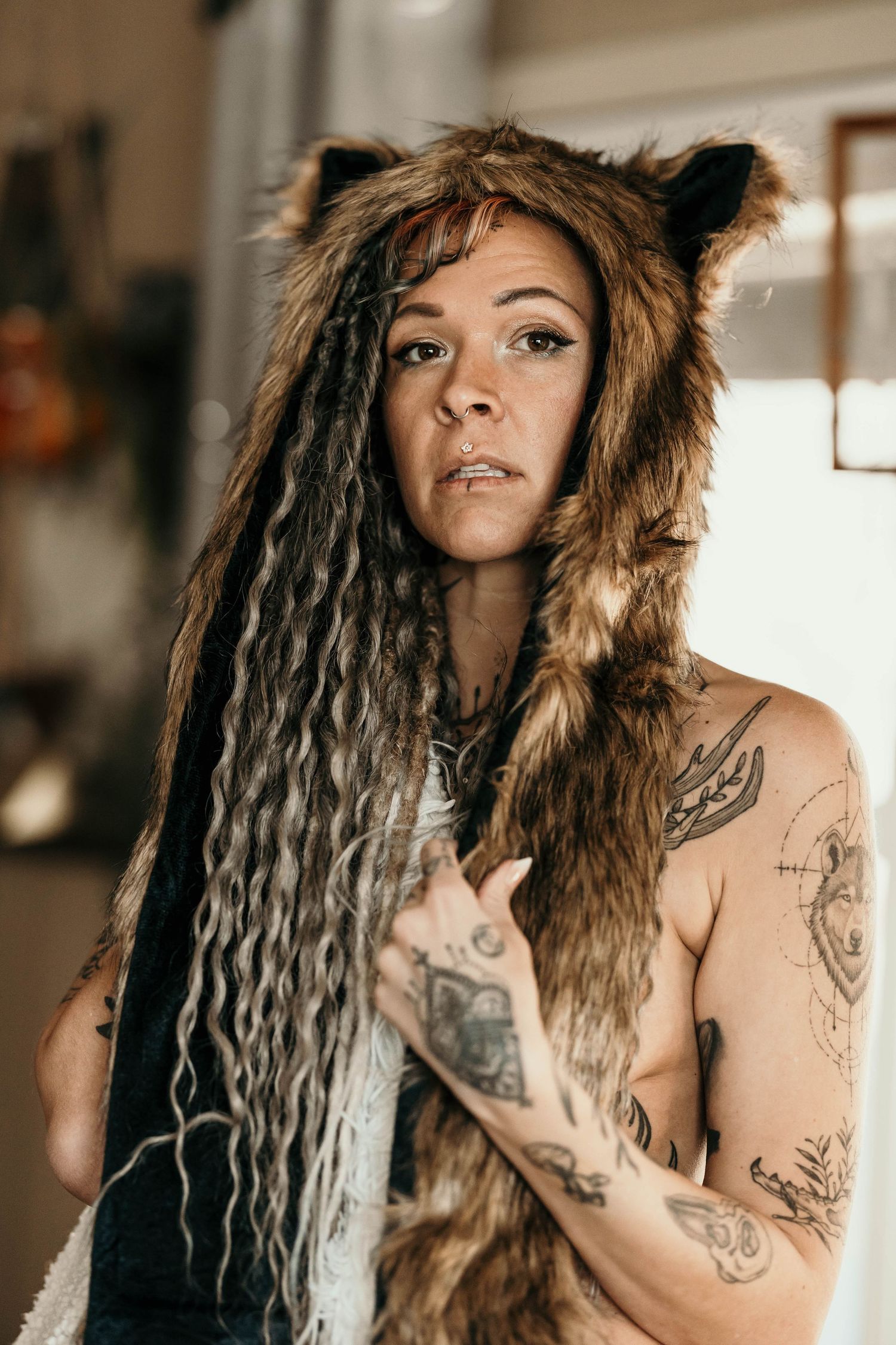 Tattooed woman wearing a fur wolf hat with long dreadlocks posing indoors with serious expression.