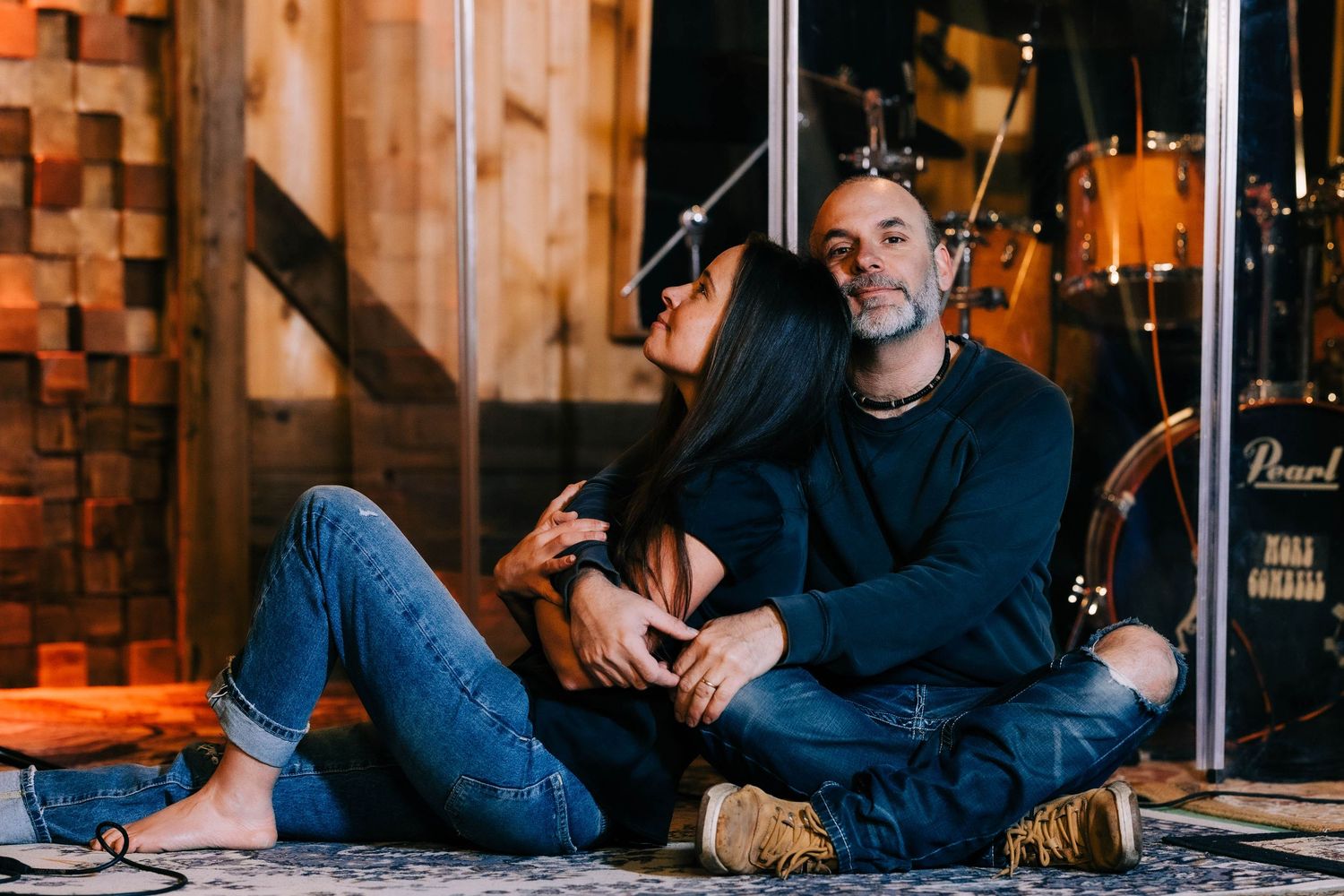 Couple sitting together on the floor in a music studio with drums visible in the background.