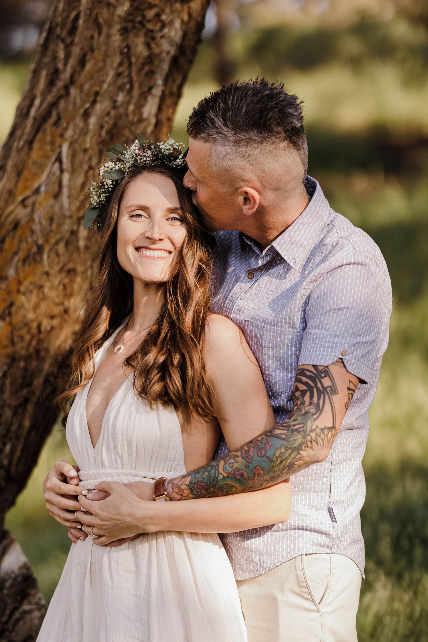 Man with tattoos kissing woman in white dress wearing floral crown by tree.