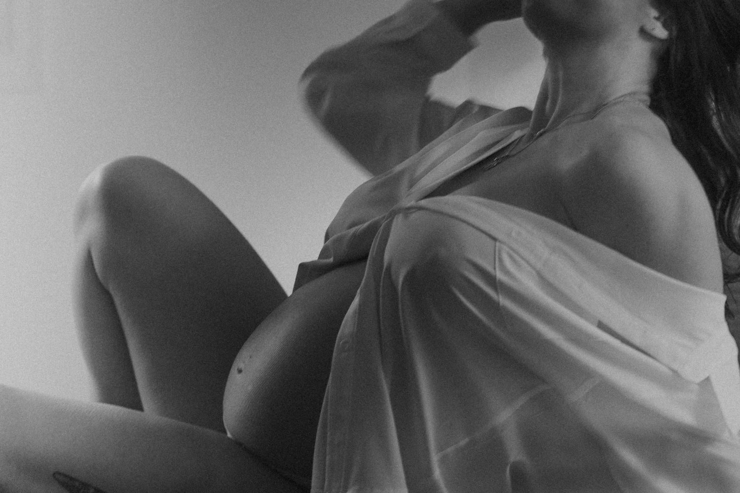 Black and white artistic photo of a pregnant woman in a sheer top sitting in soft light.