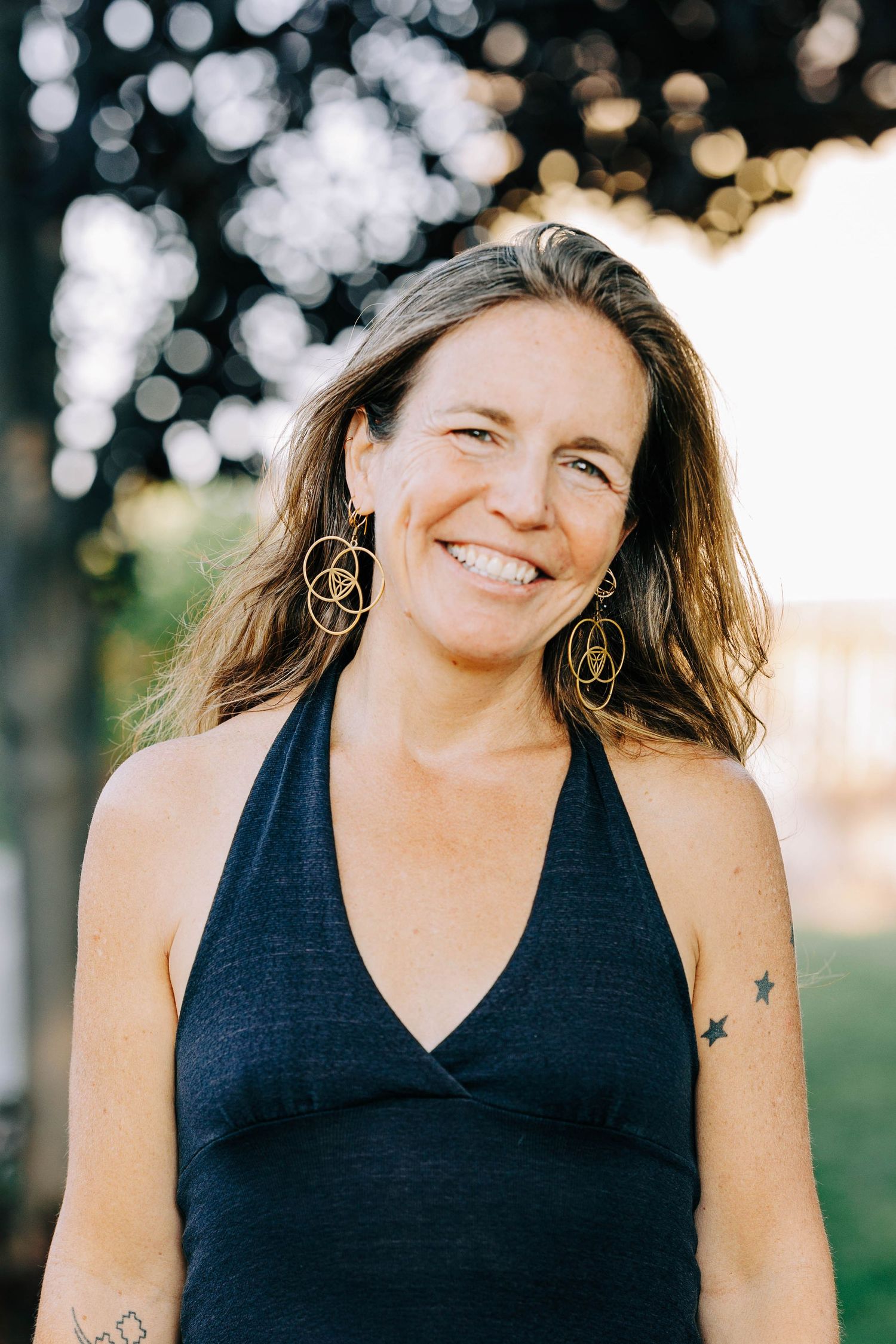 Smiling woman with long hair wearing a navy halter dress and gold earrings with visible tattoos outdoors.