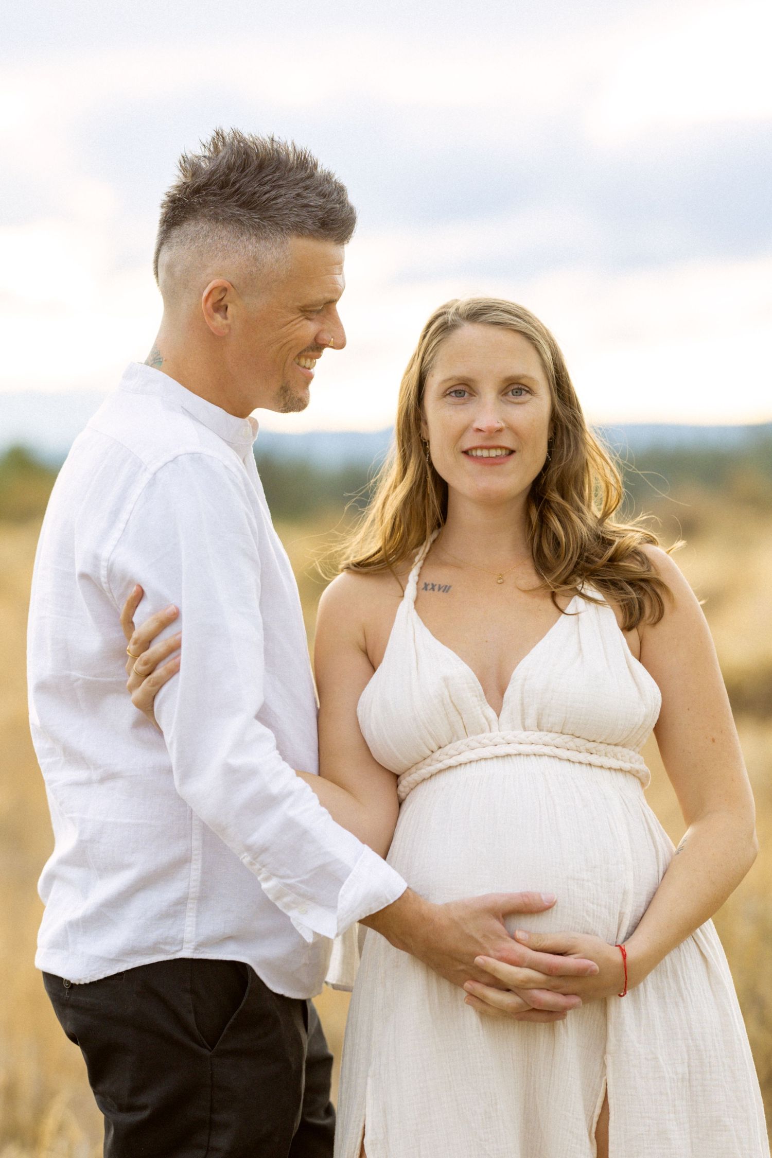 Couple posing in a golden field, man in white shirt embracing pregnant woman in white dress outdoors.