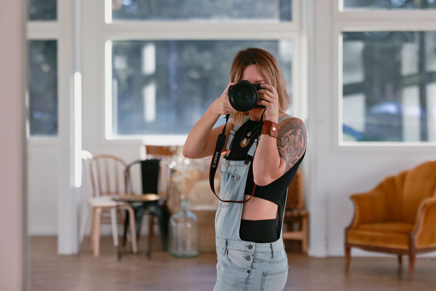 Tattooed woman photographer taking photos with DSLR camera in bright modern studio with large windows.