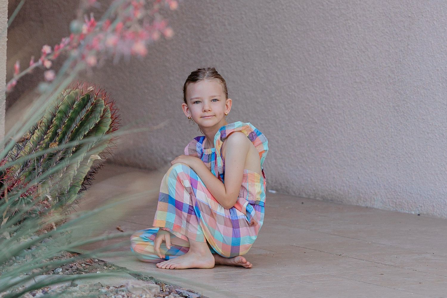 Young girl in pastel plaid jumpsuit sits cross-legged barefoot on ground beside desert plants and cacti.