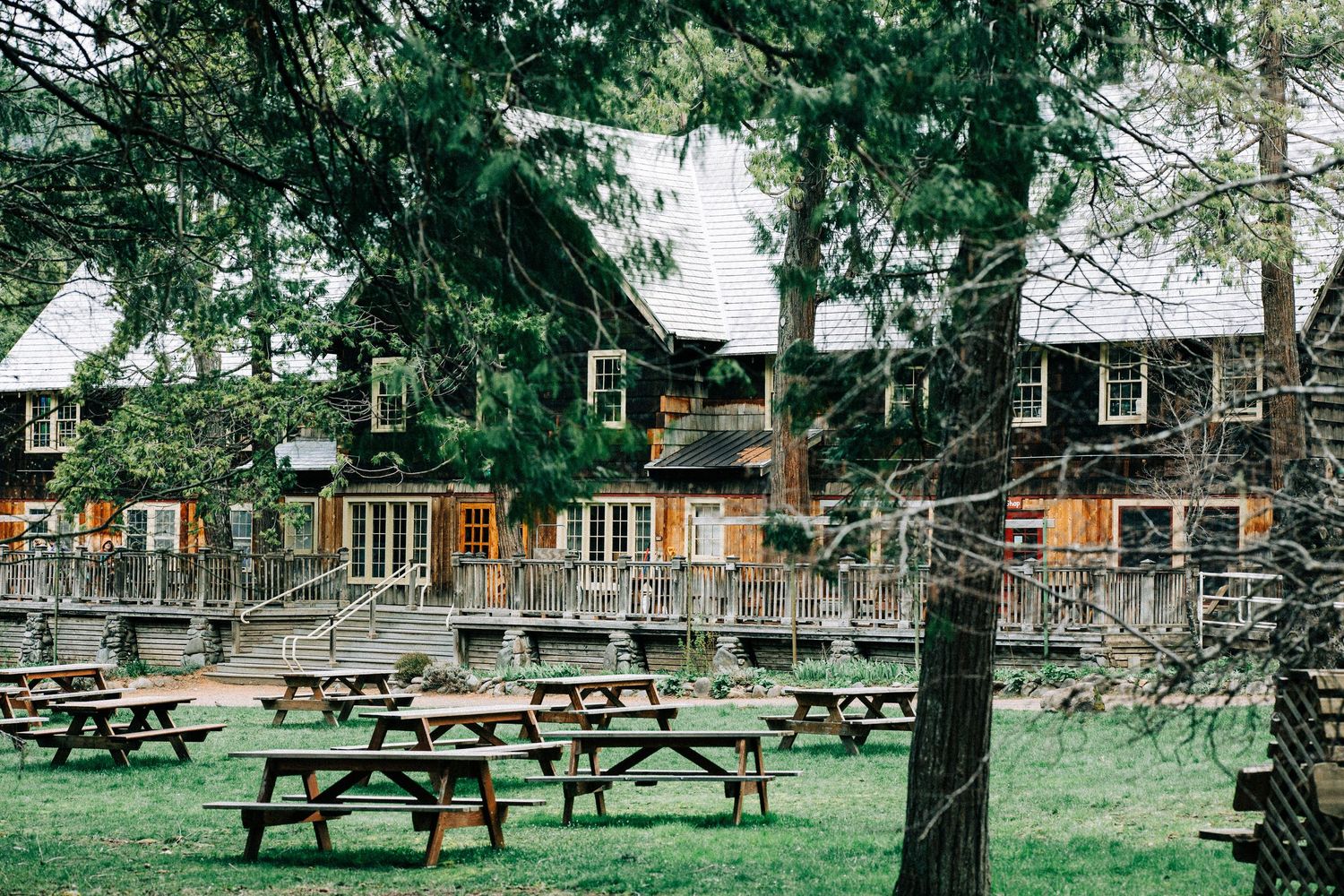 Rustic wooden lodge with picnic tables on green lawn under trees.