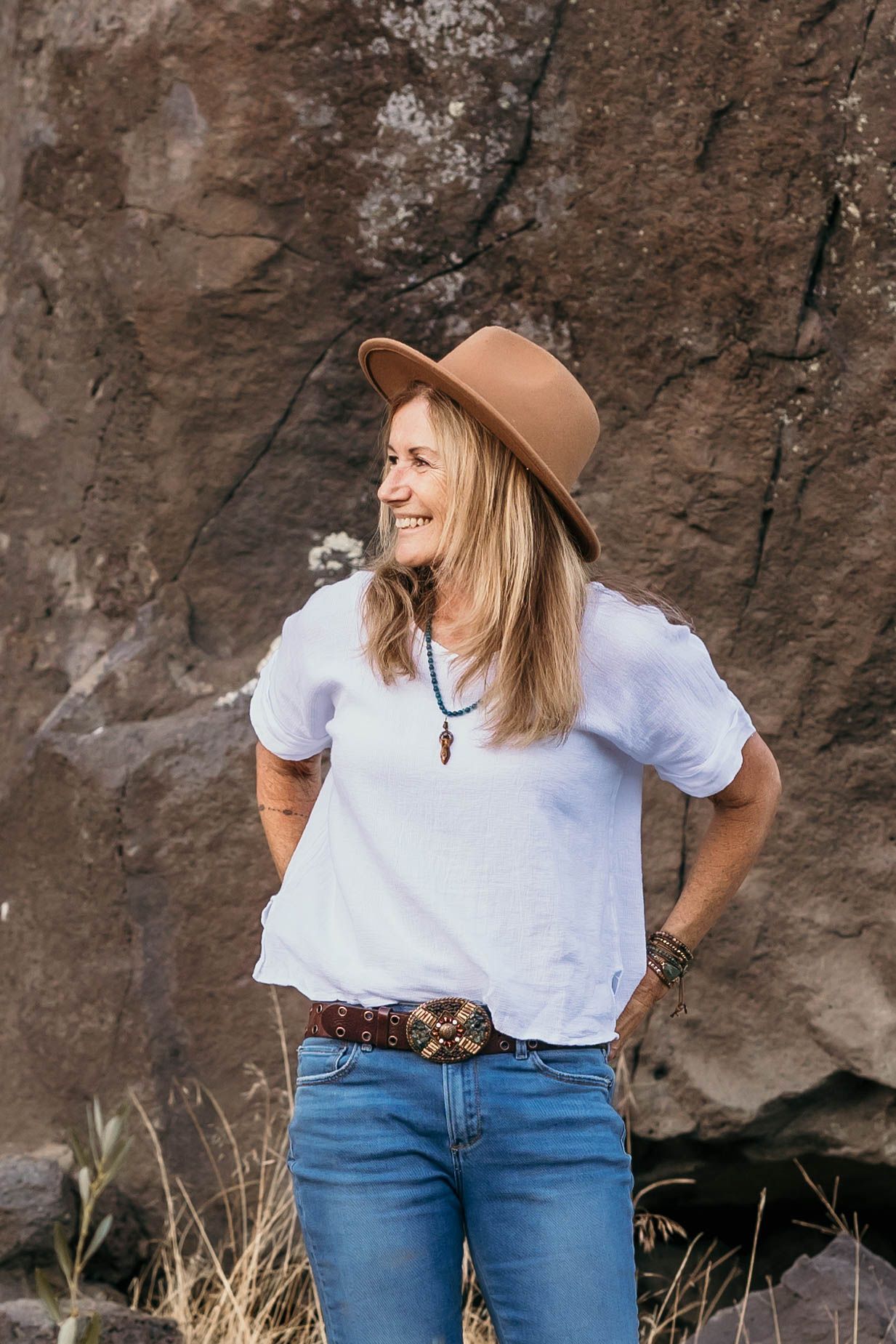 Blonde woman in white tee, jeans, and brown hat by a rock.