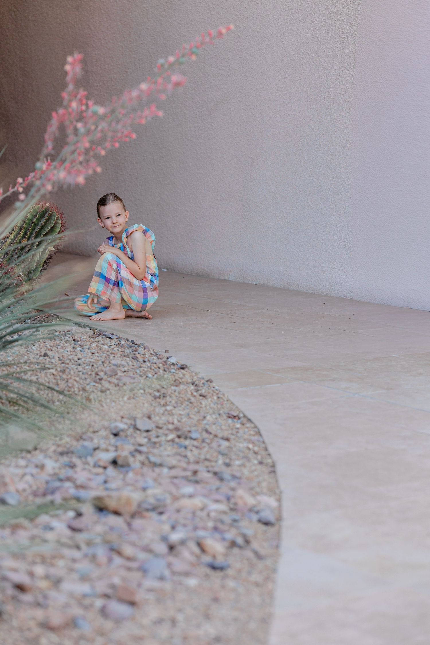 Young girl in colorful plaid dress sitting on patio near desert garden plants.