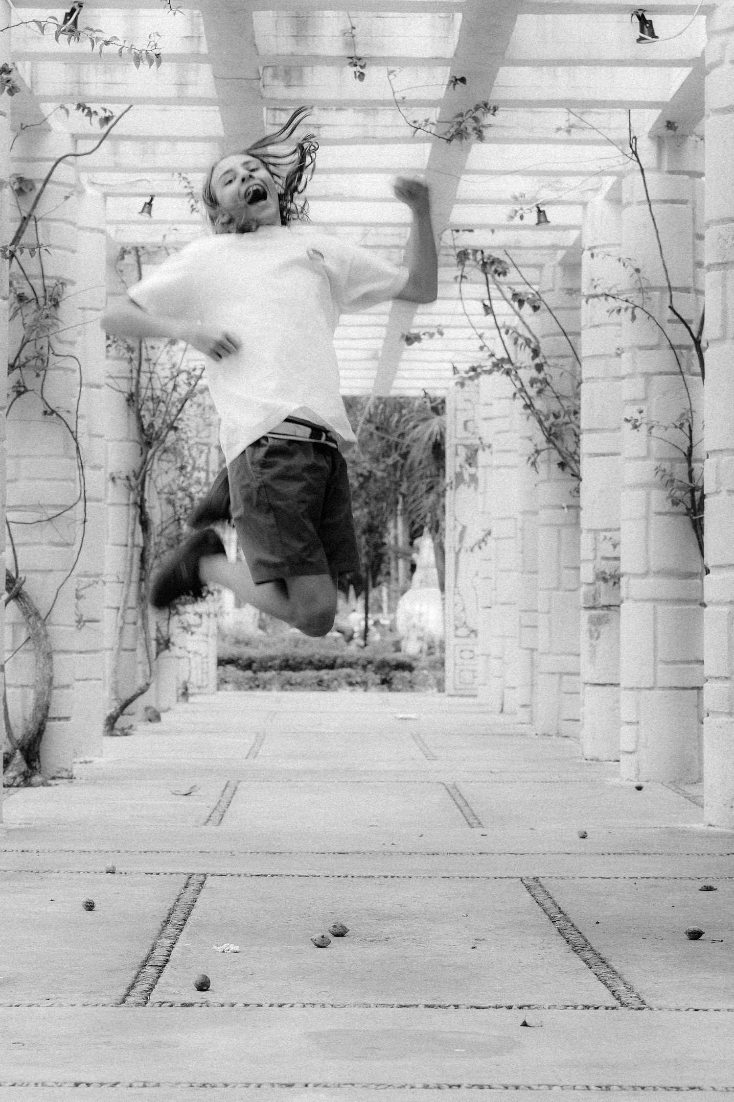Black and white photo of an energetic boy jumping mid-air in an outdoor pergola corridor.