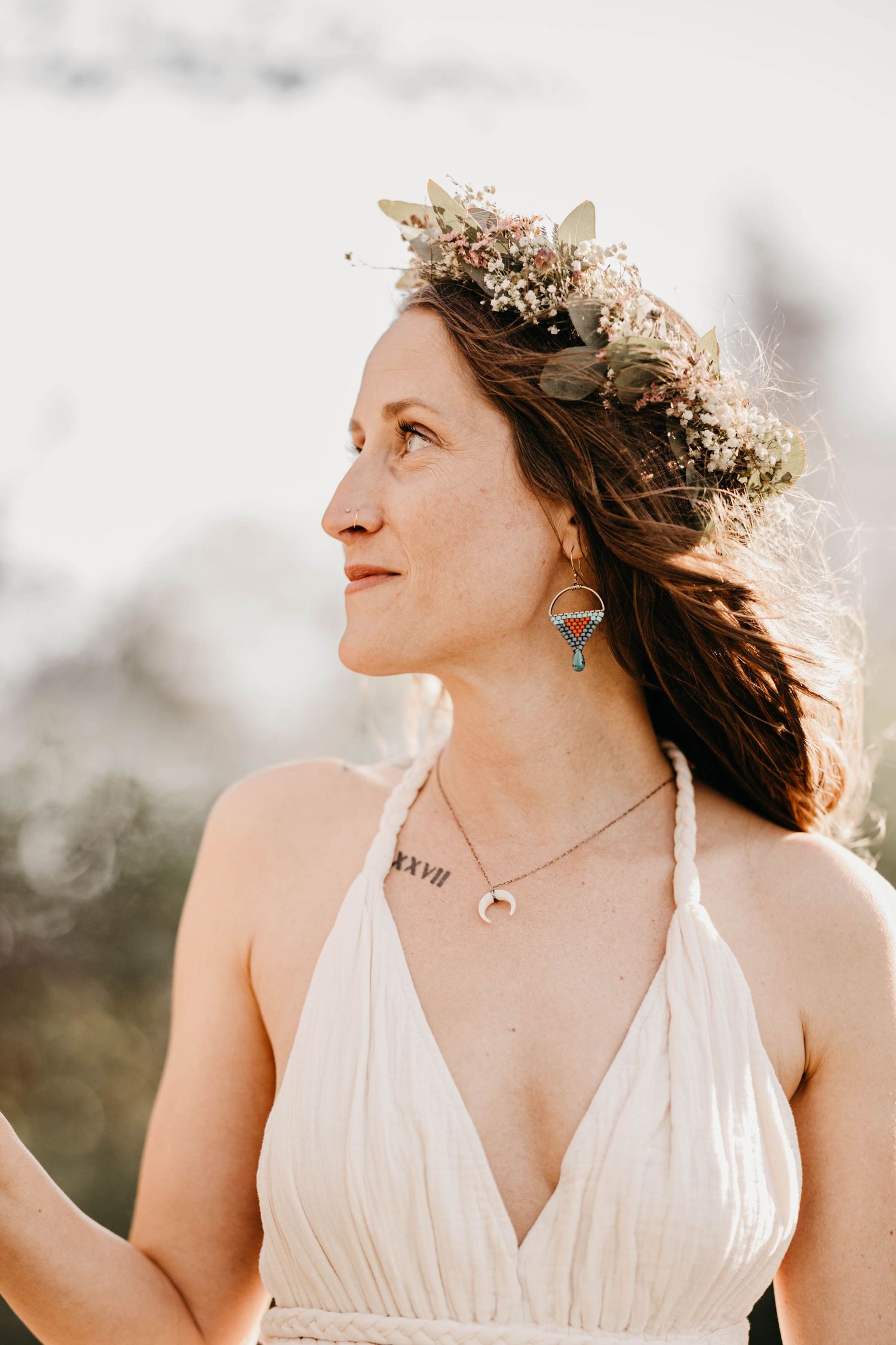 Woman wearing a floral crown and white halter dress with crescent moon necklace looking sideways outdoors.