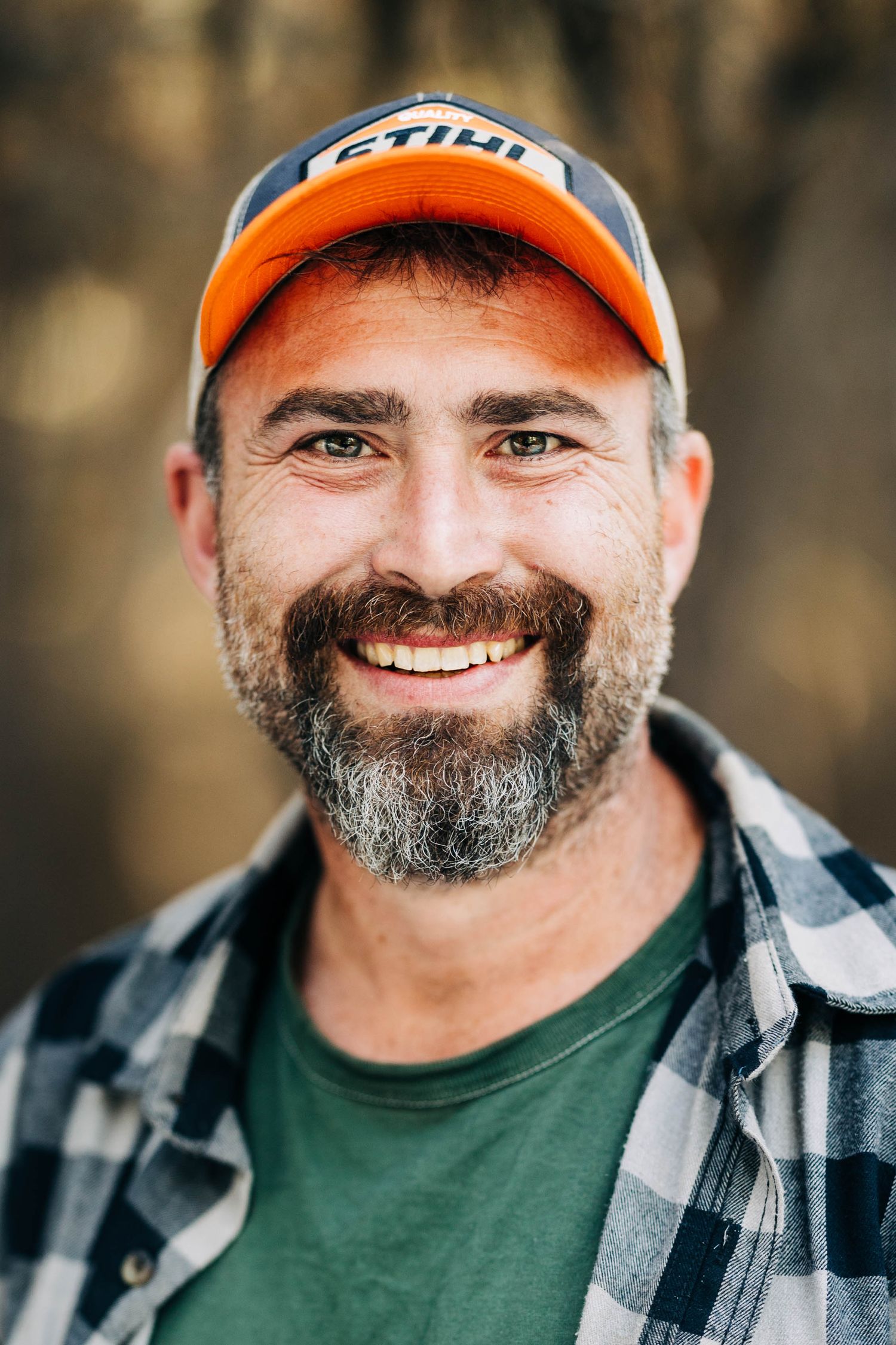 Bearded man smiling in plaid shirt and orange cap outdoors.