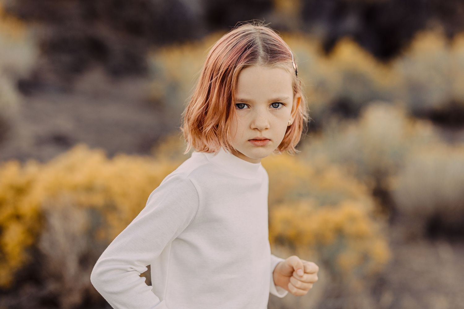 Young girl with reddish-blonde bob haircut wearing white sweater poses outdoors with golden foliage background.
