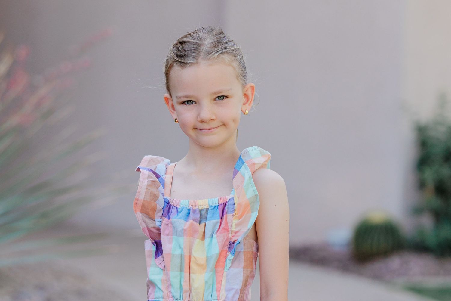 Cute young girl in colorful pastel plaid romper smiles sweetly while standing outdoors near cacti.