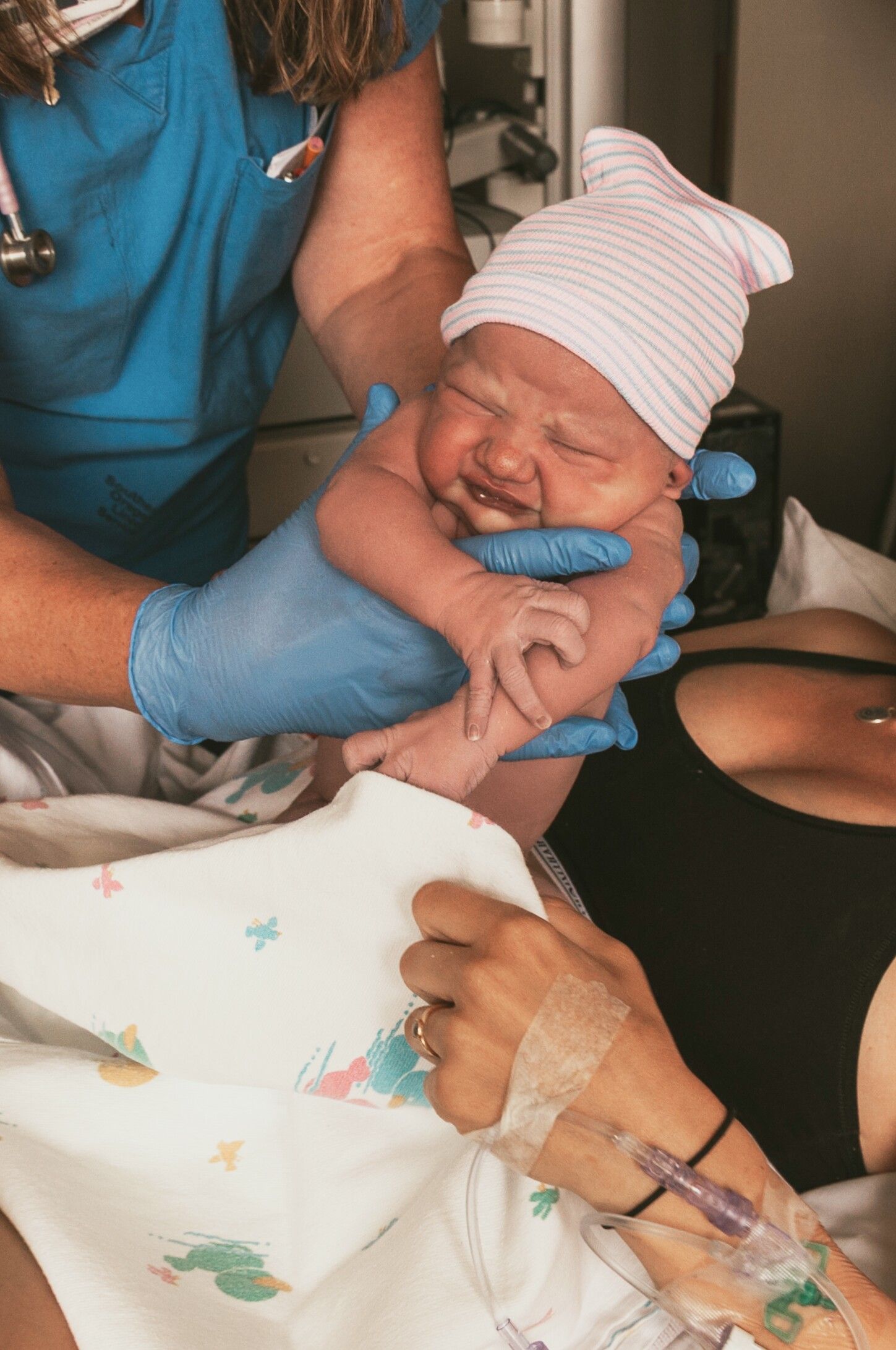 Nurse in blue scrubs presenting alert newborn with pink hat to mother.