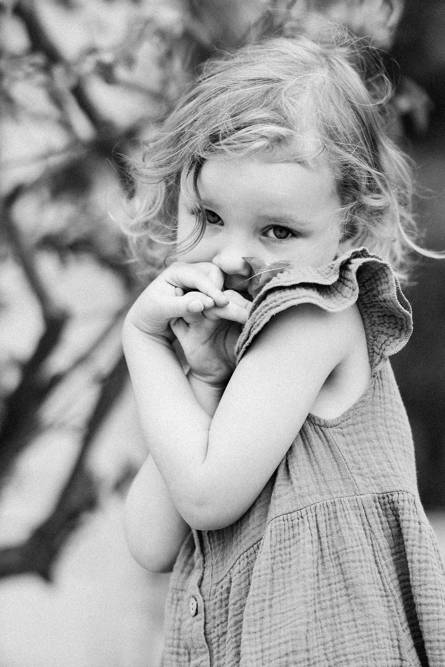 Black and white portrait of a curly-haired toddler girl holding her dress near her face.