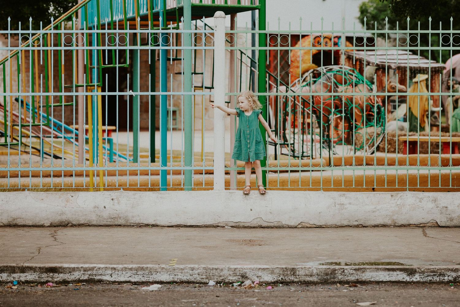 A young girl in a teal dress stands by a colorful playground fence on a sunny day.