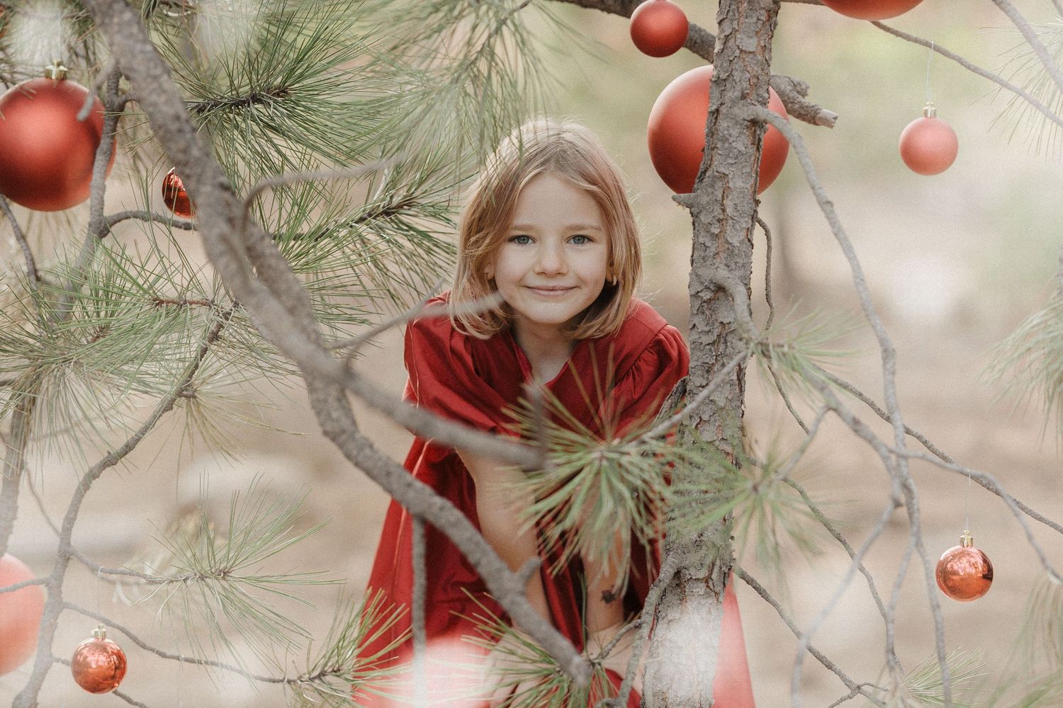 A smiling girl in a red velvet dress poses among pine branches decorated with red Christmas ornaments.