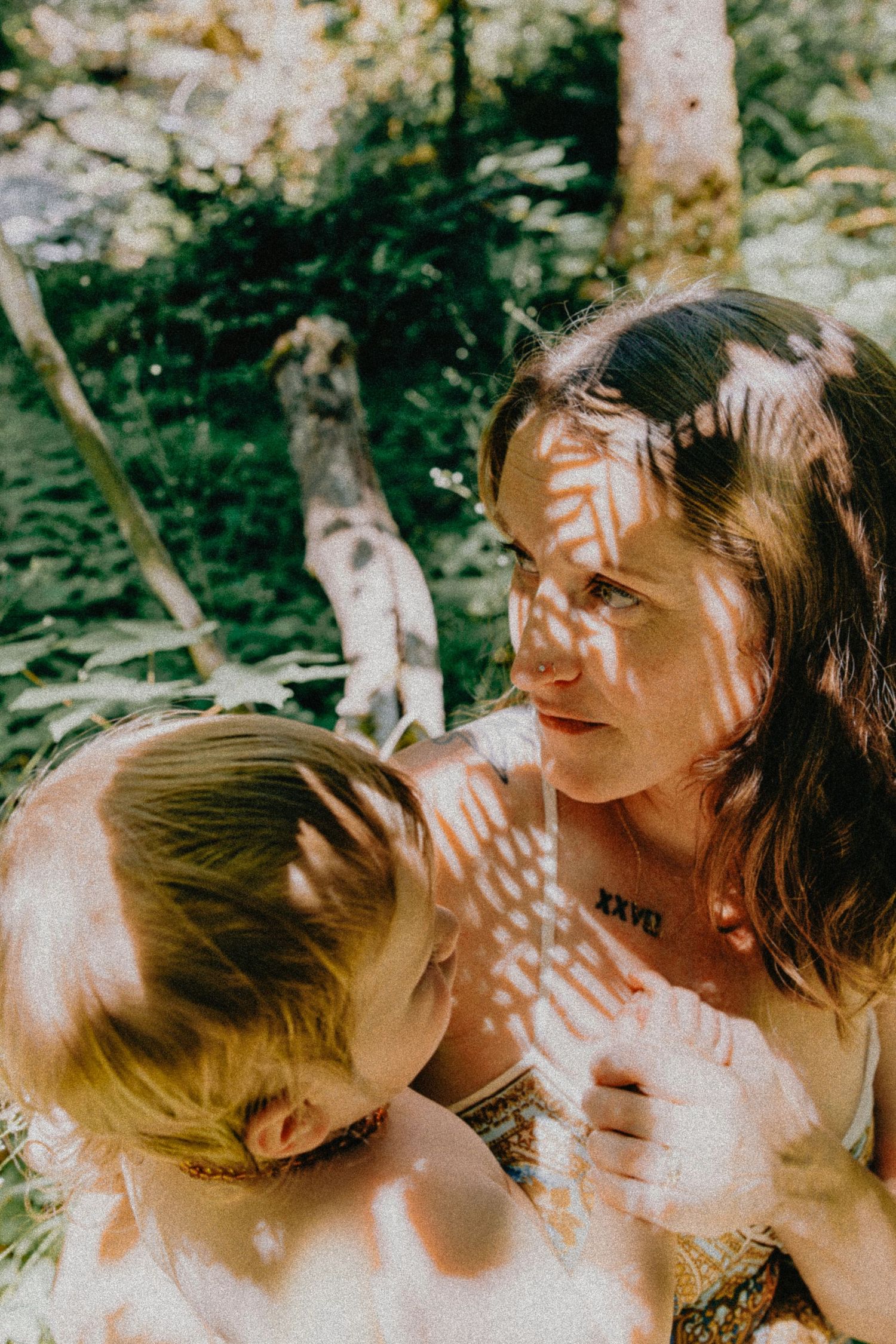 Mother and young child sharing a tender moment in a sunlit forest with dappled shadows.