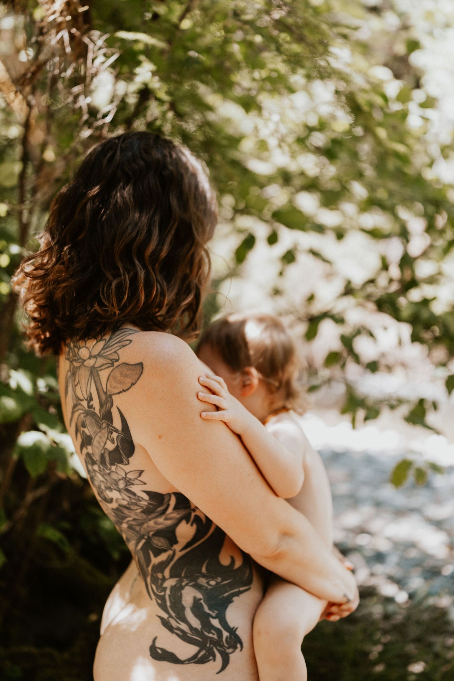 Mother with an intricate back tattoo holding her young child outdoors in natural light.