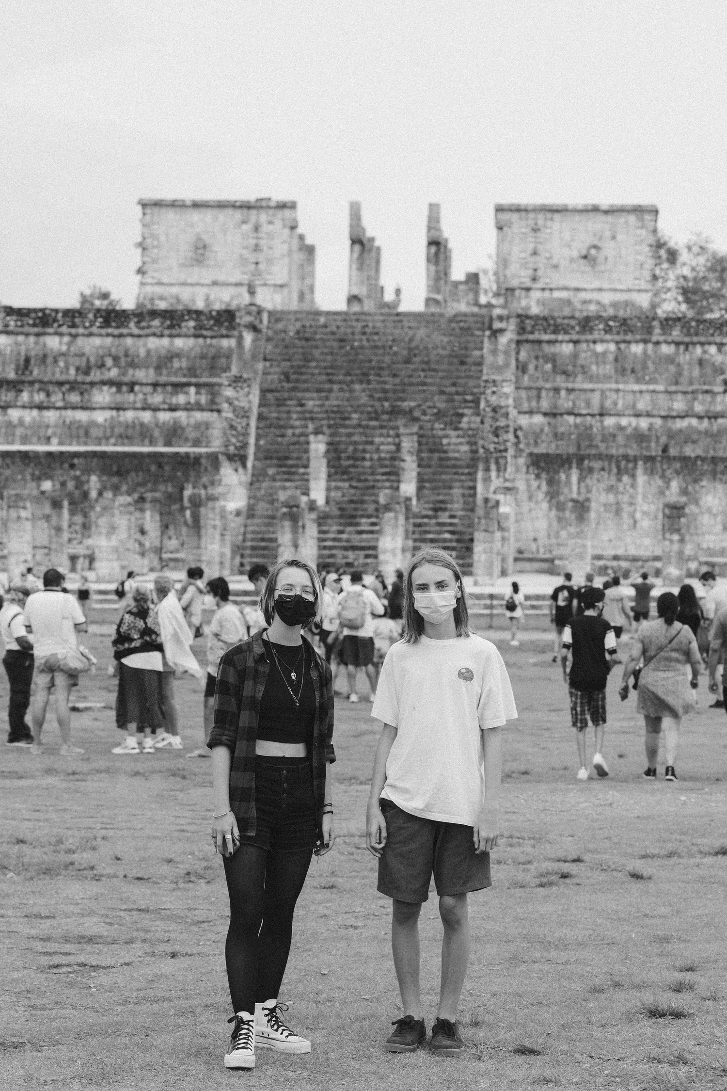 Two masked tourists in black and white photo standing before Chichen Itza ruins, Mexico.