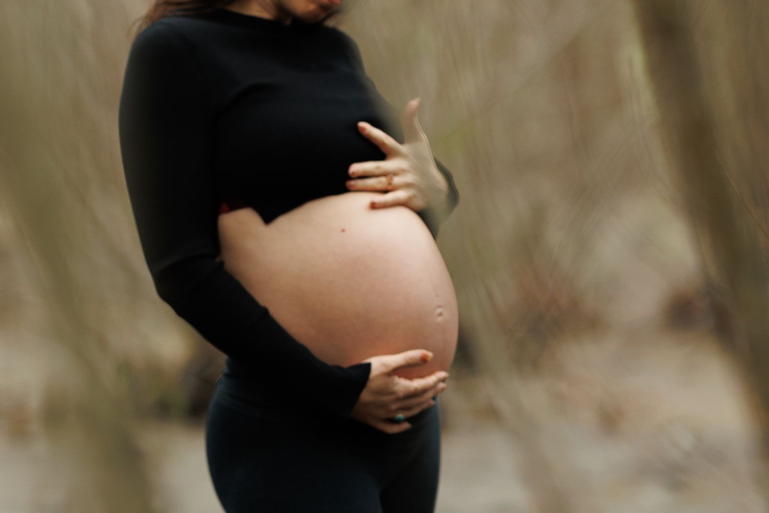 Close-up of pregnant woman in black outfit cradling her bare baby bump in a forest setting.