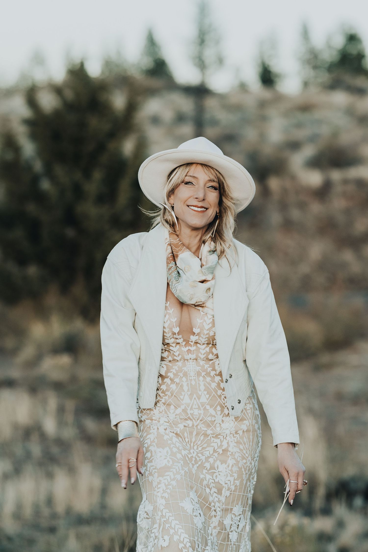 Smiling blonde woman in white hat and lace dress laughing outdoors in natural setting.