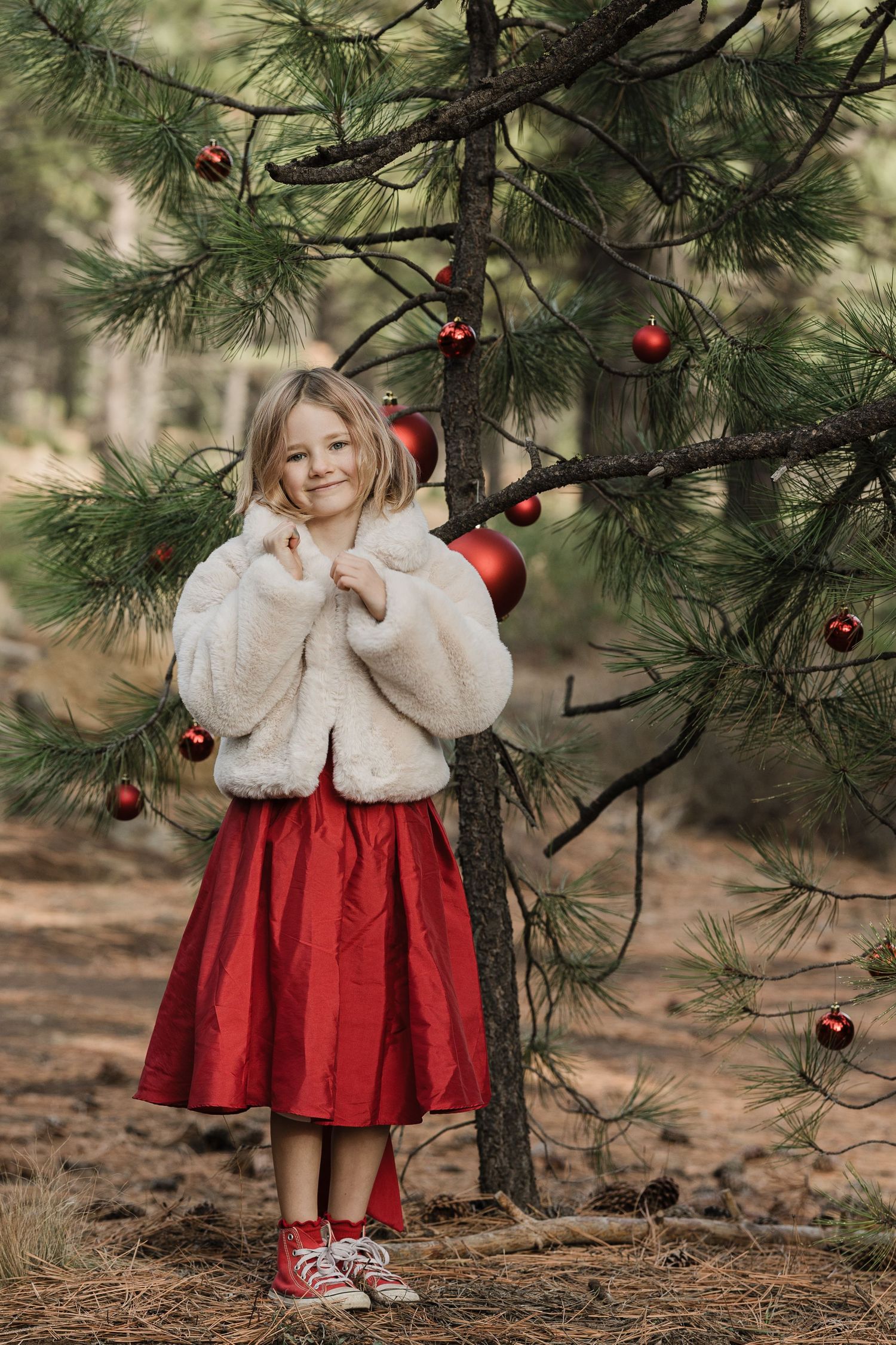 Young girl in white fur jacket and red dress standing beside pine tree with red Christmas ornaments.