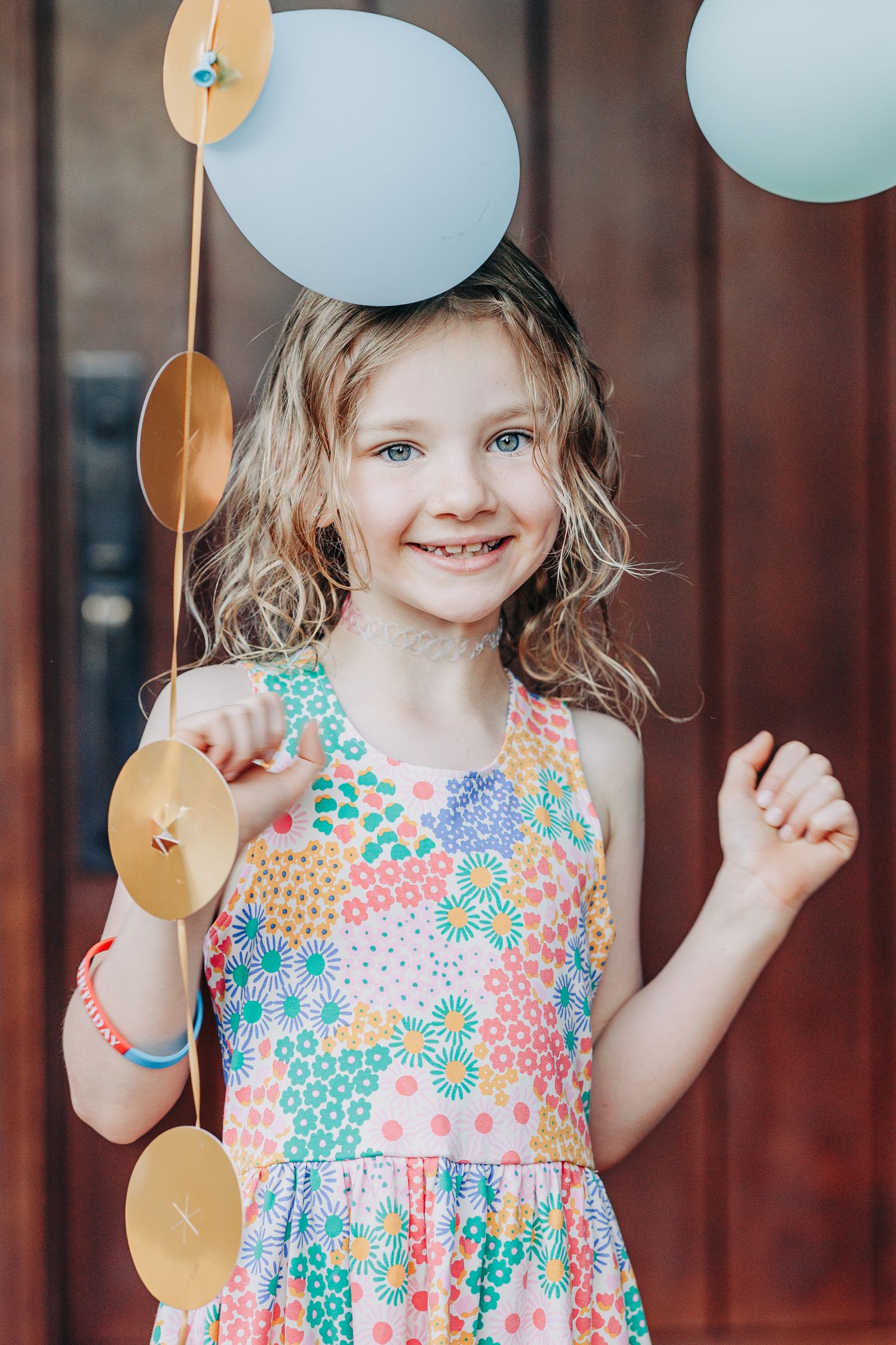 Happy young girl in colorful floral dress smiling while playing with decorative balloon garland.