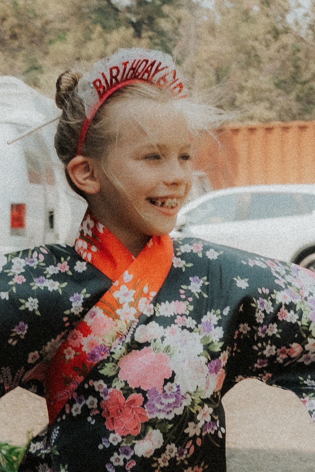 Young girl in floral dress wearing birthday girl tiara laughing outdoors.