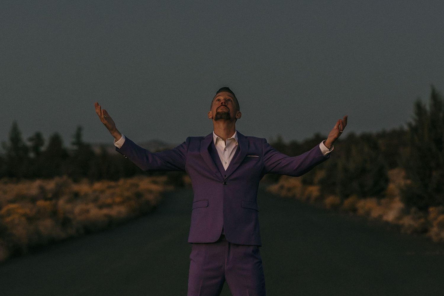 Man in purple suit standing on road with arms outstretched under dramatic evening sky.