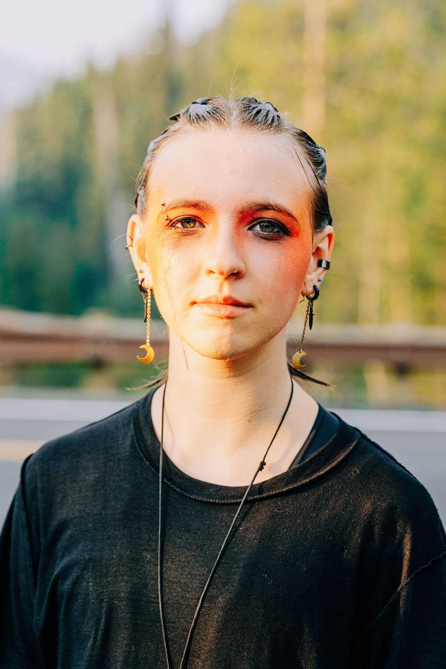 Young woman with bold red eye makeup and moon earrings standing outdoors with forest and road in background.