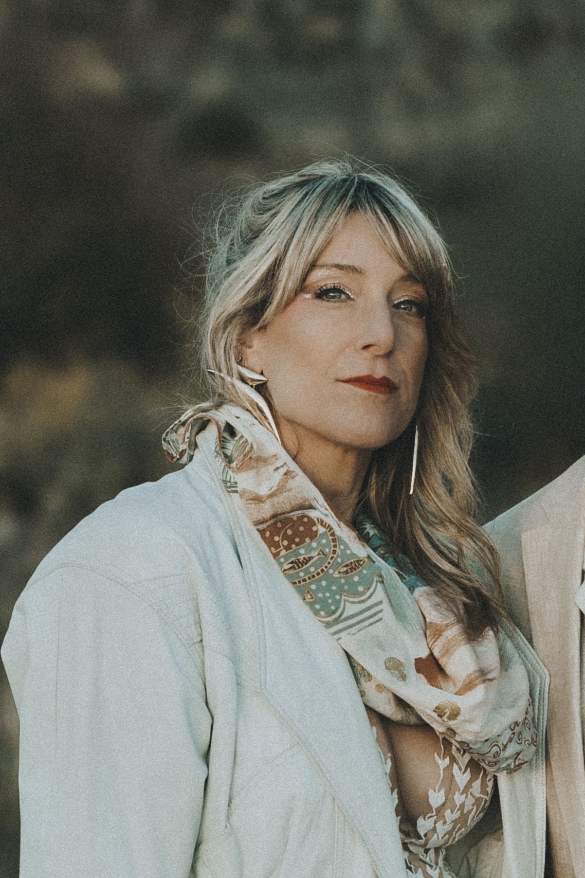Blonde woman in white jacket and floral scarf posing outdoors with soft natural background.