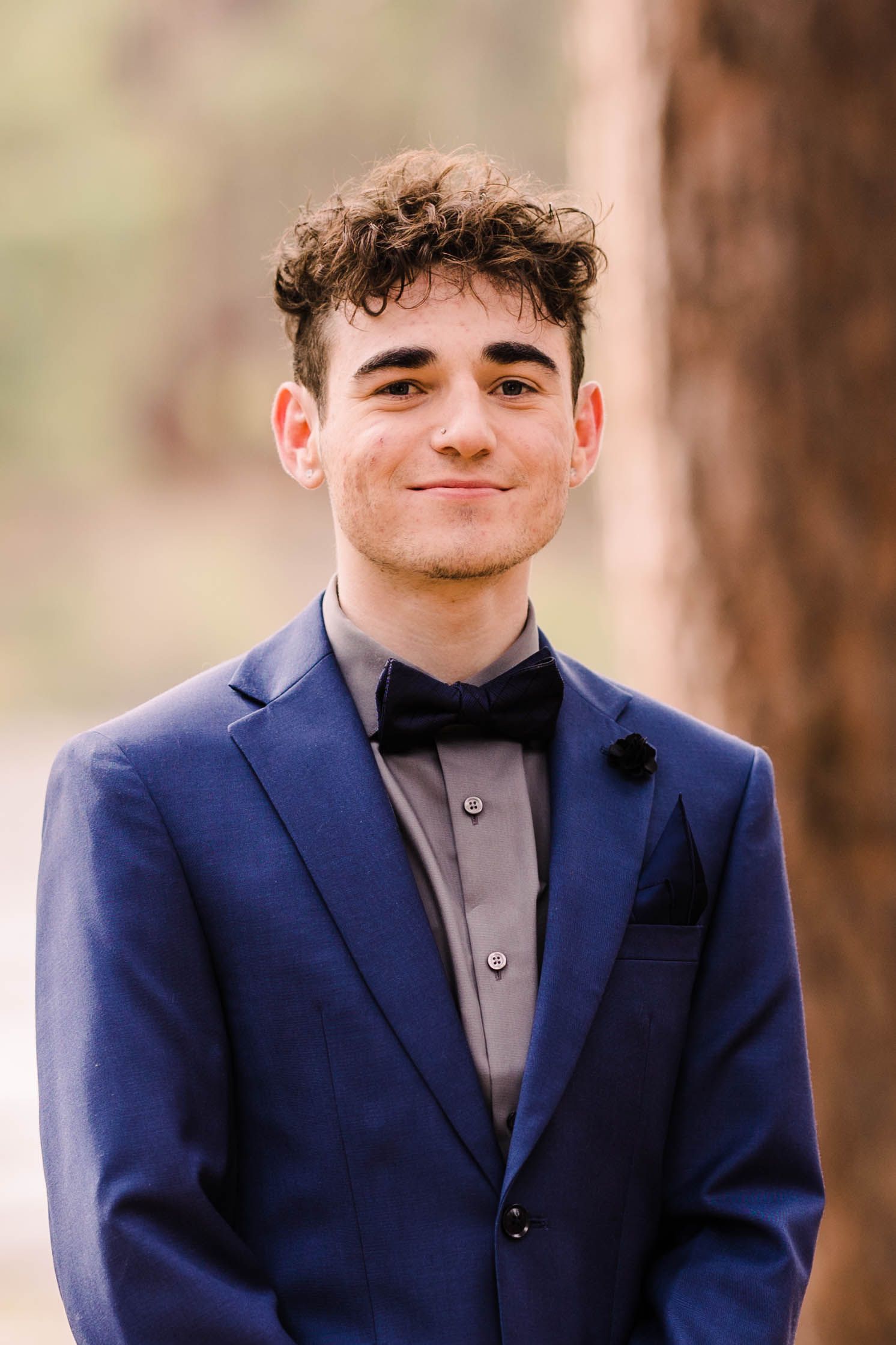 Smiling young man in blue blazer with black bow tie and curly hair posing near a tree outdoors.