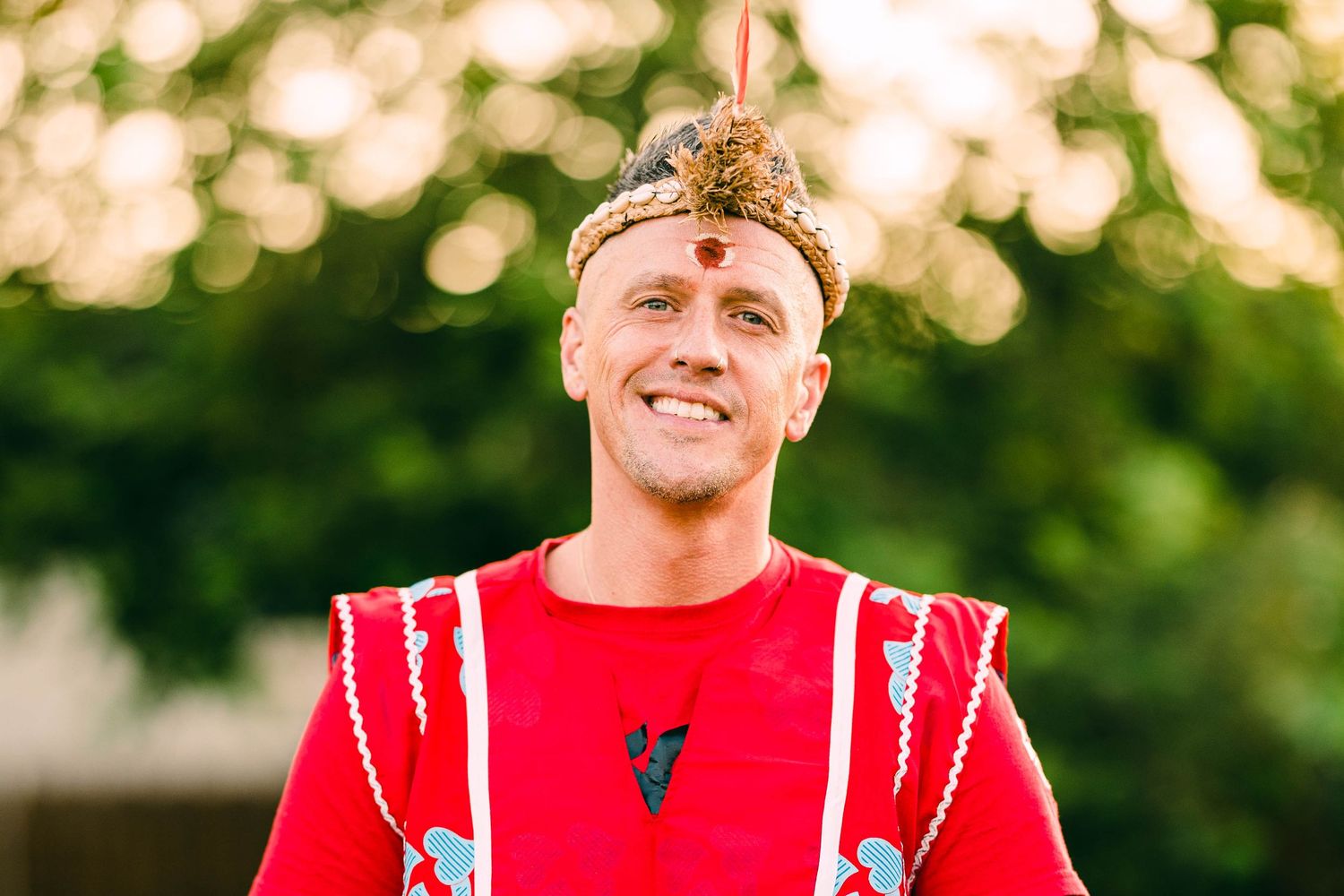 Smiling man wearing a red traditional ceremonial outfit with decorative headpiece outdoors in sunlight.