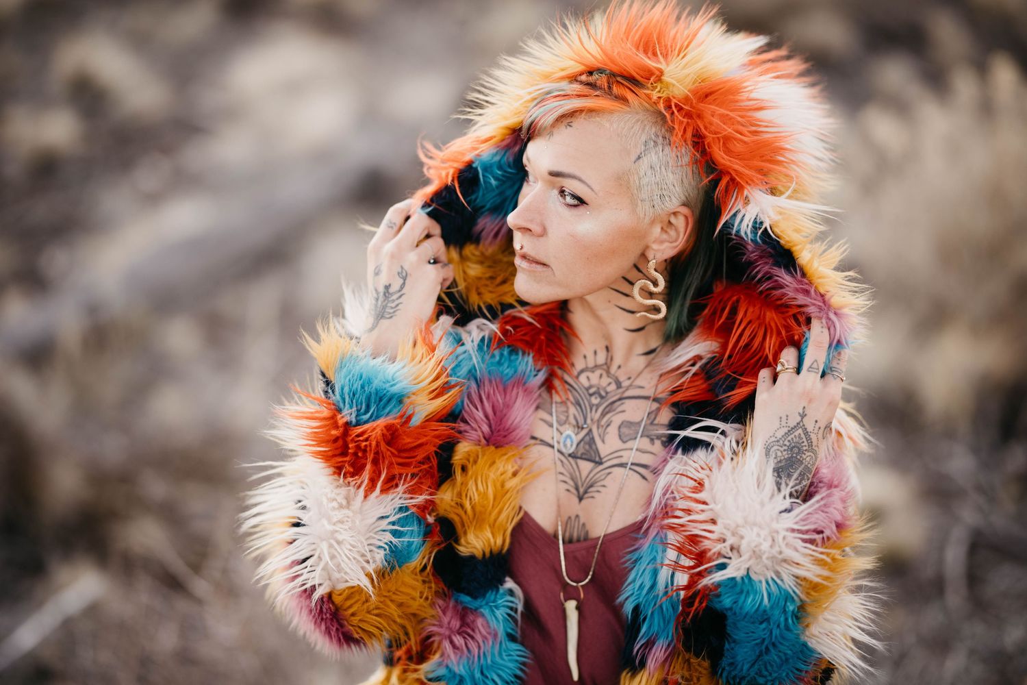 Tattooed woman in vibrant multicolor faux fur coat poses in dry outdoor landscape.