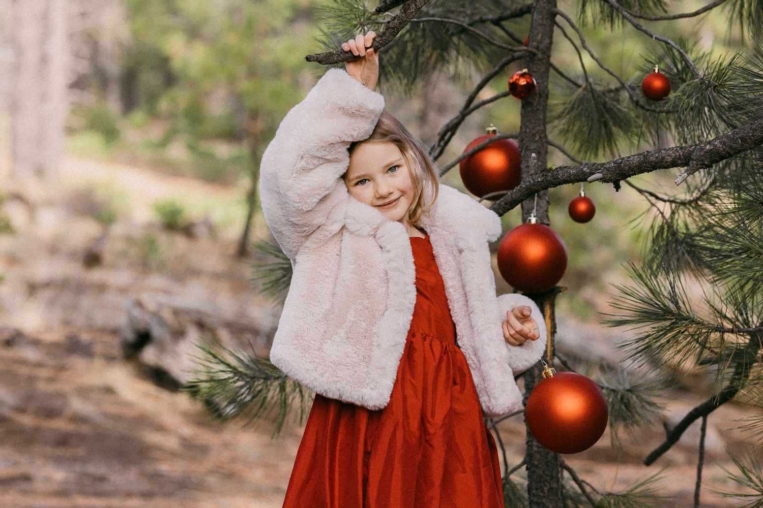 Young girl in red dress and white fur jacket posing by pine tree decorated with red Christmas ornaments.
