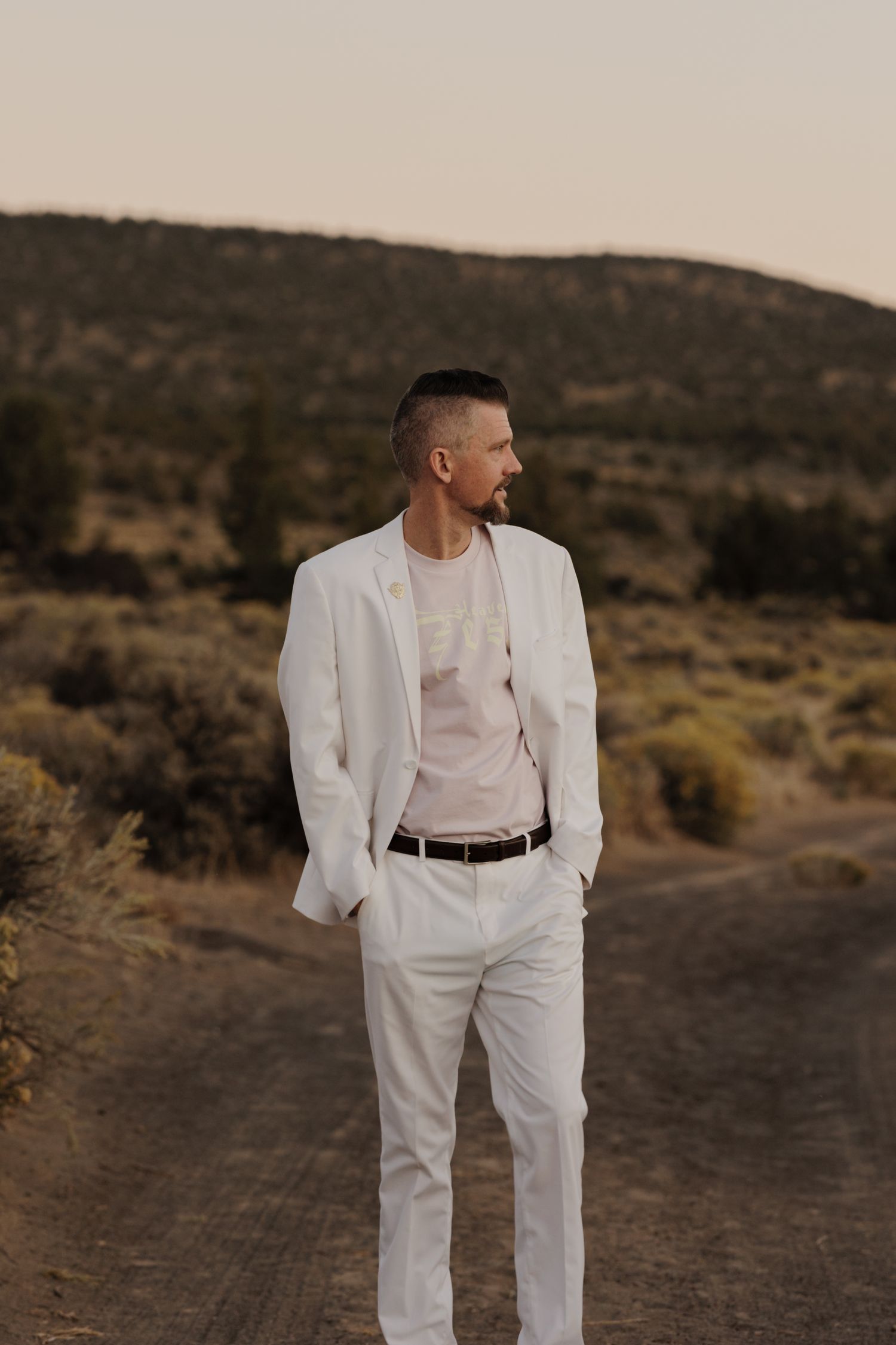 Man in white suit and t-shirt standing on dirt road with sagebrush hills at dusk.